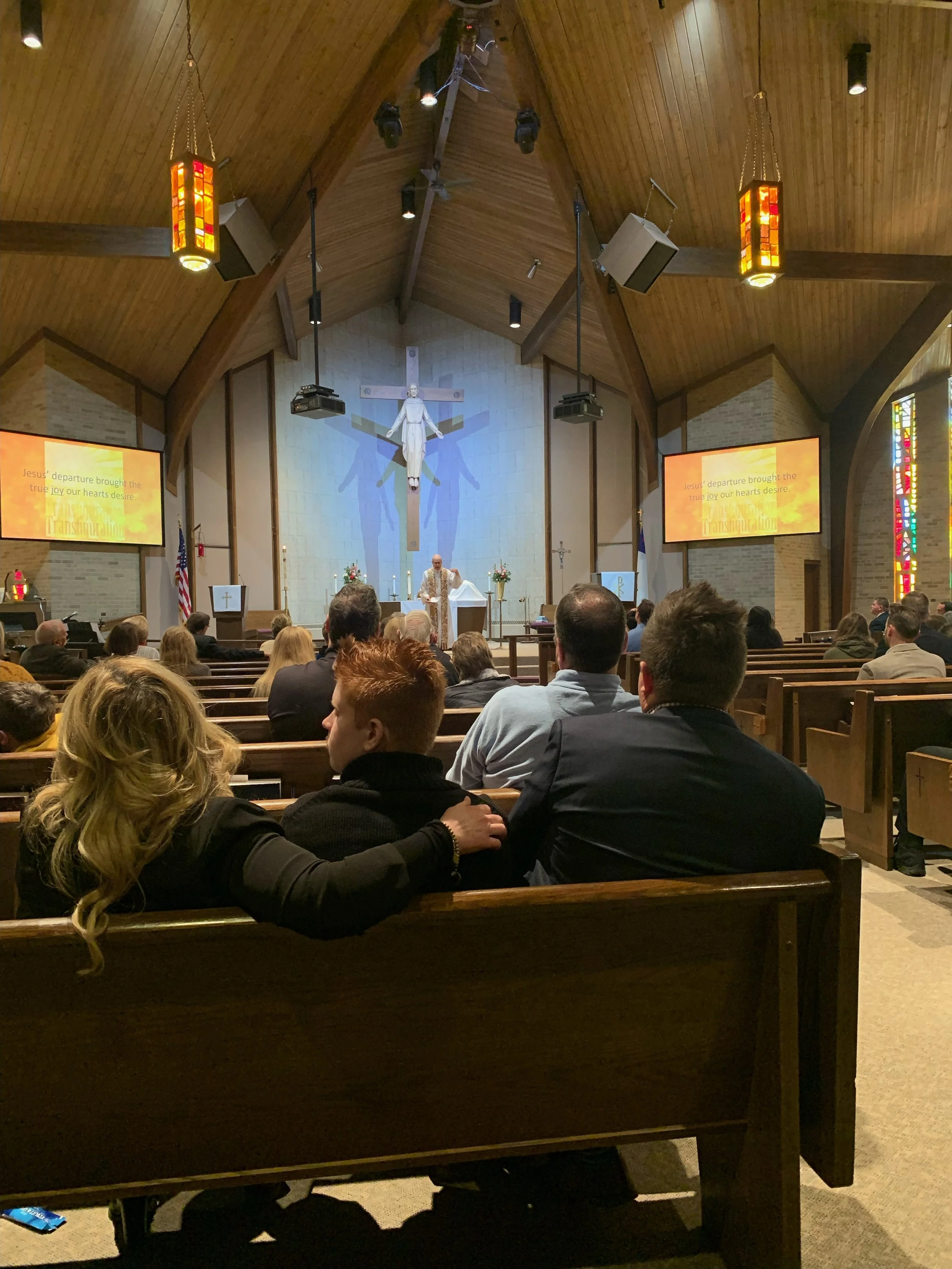 Inside a church with a congregation sitting on wooden pews, facing the altar. The altar features a large cross with a crucifix of Jesus. The church has stained glass windows, hanging lamps, and two screens on either side of the altar displaying lyrics.