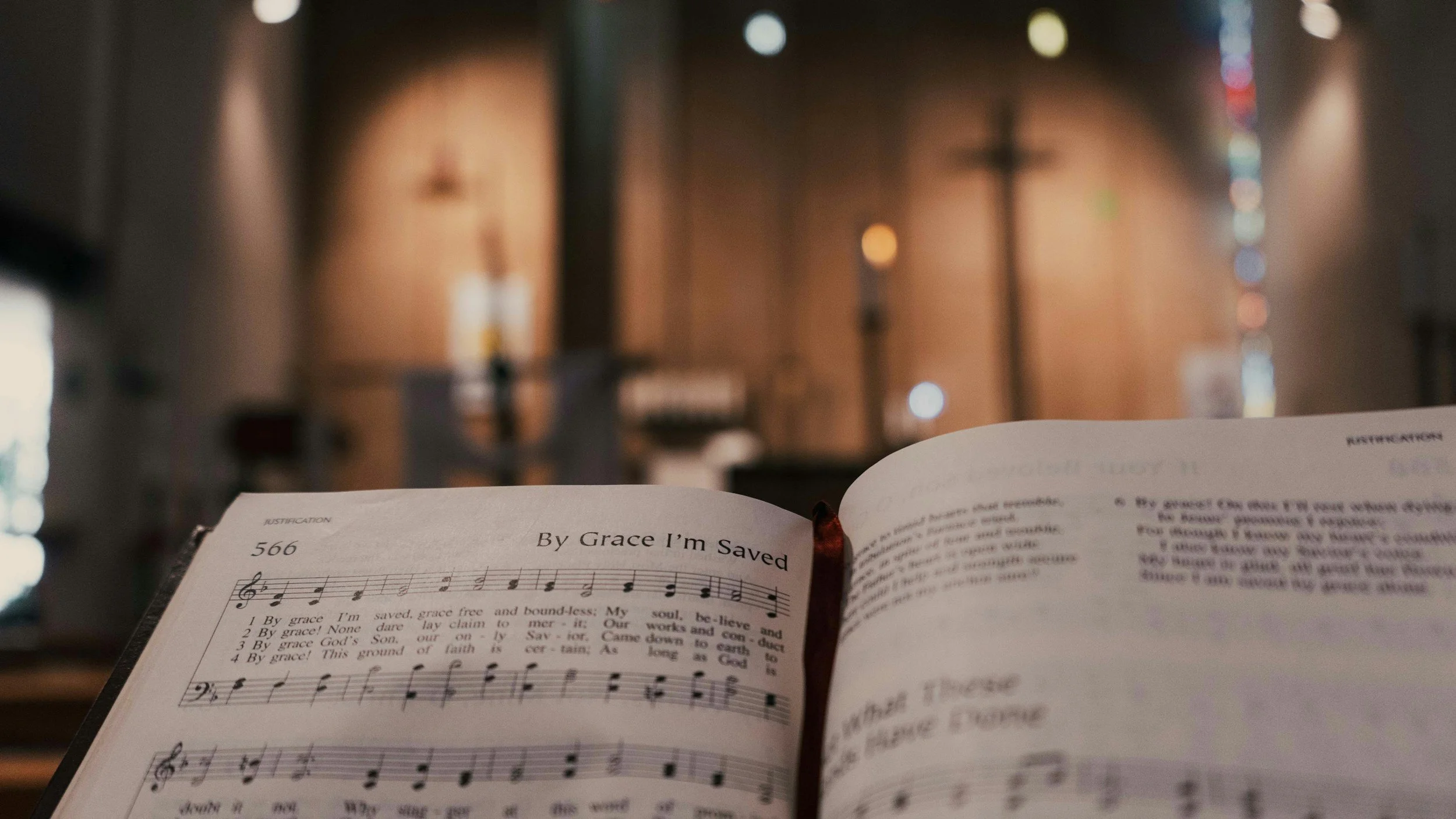 Open hymnal with sheet music titled "By Grace I'm Saved" inside a church with blurred cross and stained glass on the wall in the background.