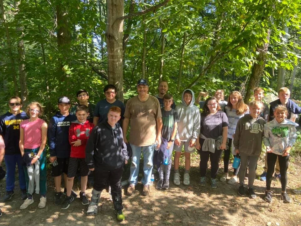 Group of children and adults standing together outdoors in a forest, smiling for the camera.