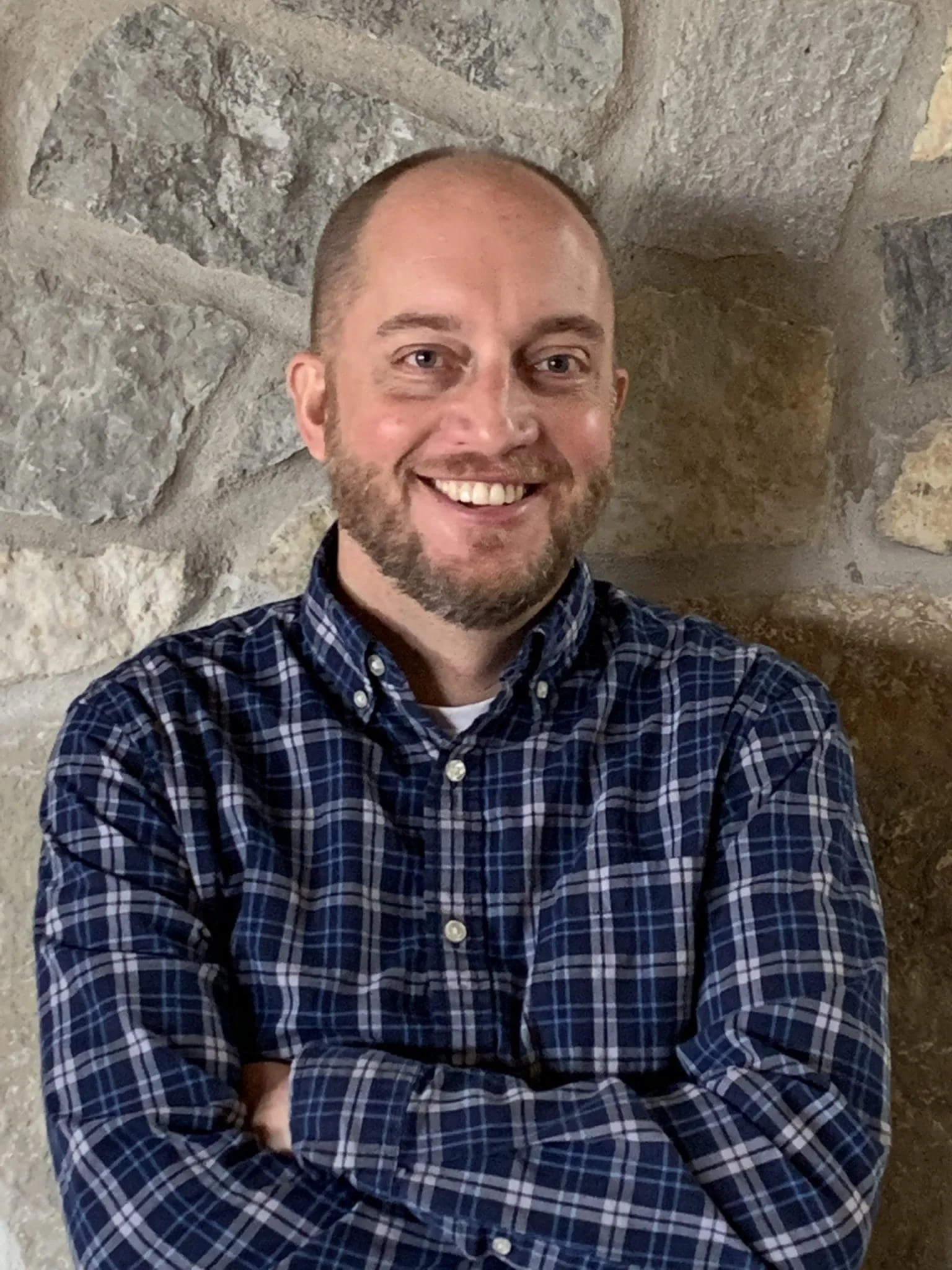 A smiling man with a beard and short hair, wearing a blue plaid shirt, stands in front of a stone wall with crossed arms.