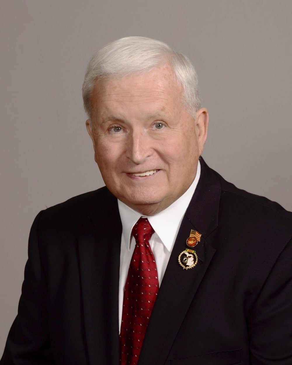A senior man with white hair, wearing a black suit, white shirt, and red tie, smiling at the camera. He has pins on his suit lapel.