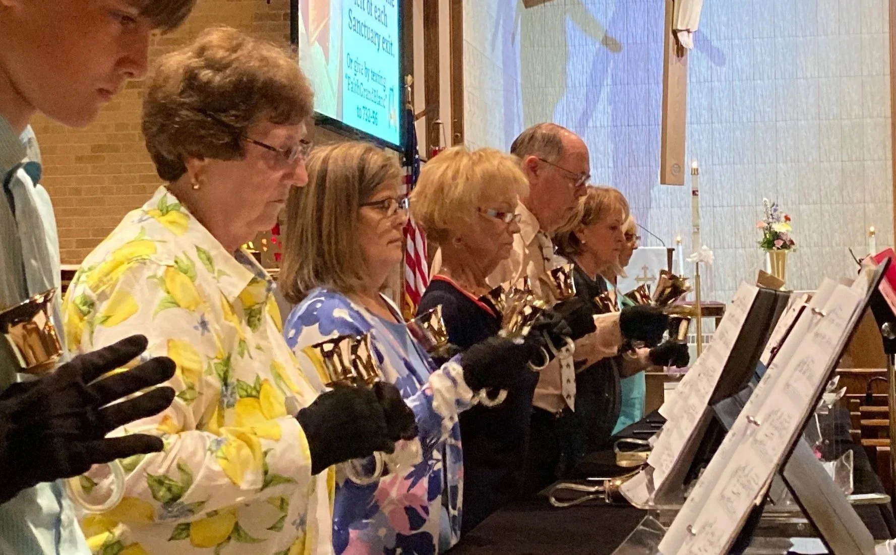A group of people standing in a church holding handbells, participating in a prayer or moment of reflection during a religious service.
