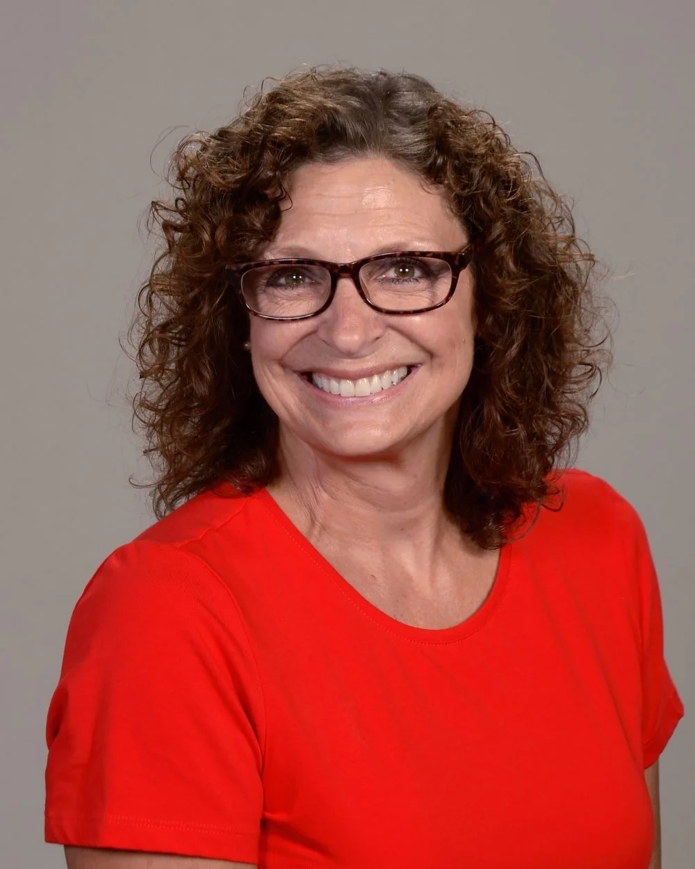 A woman with curly brown hair and glasses smiling, wearing a red shirt against a plain gray background.