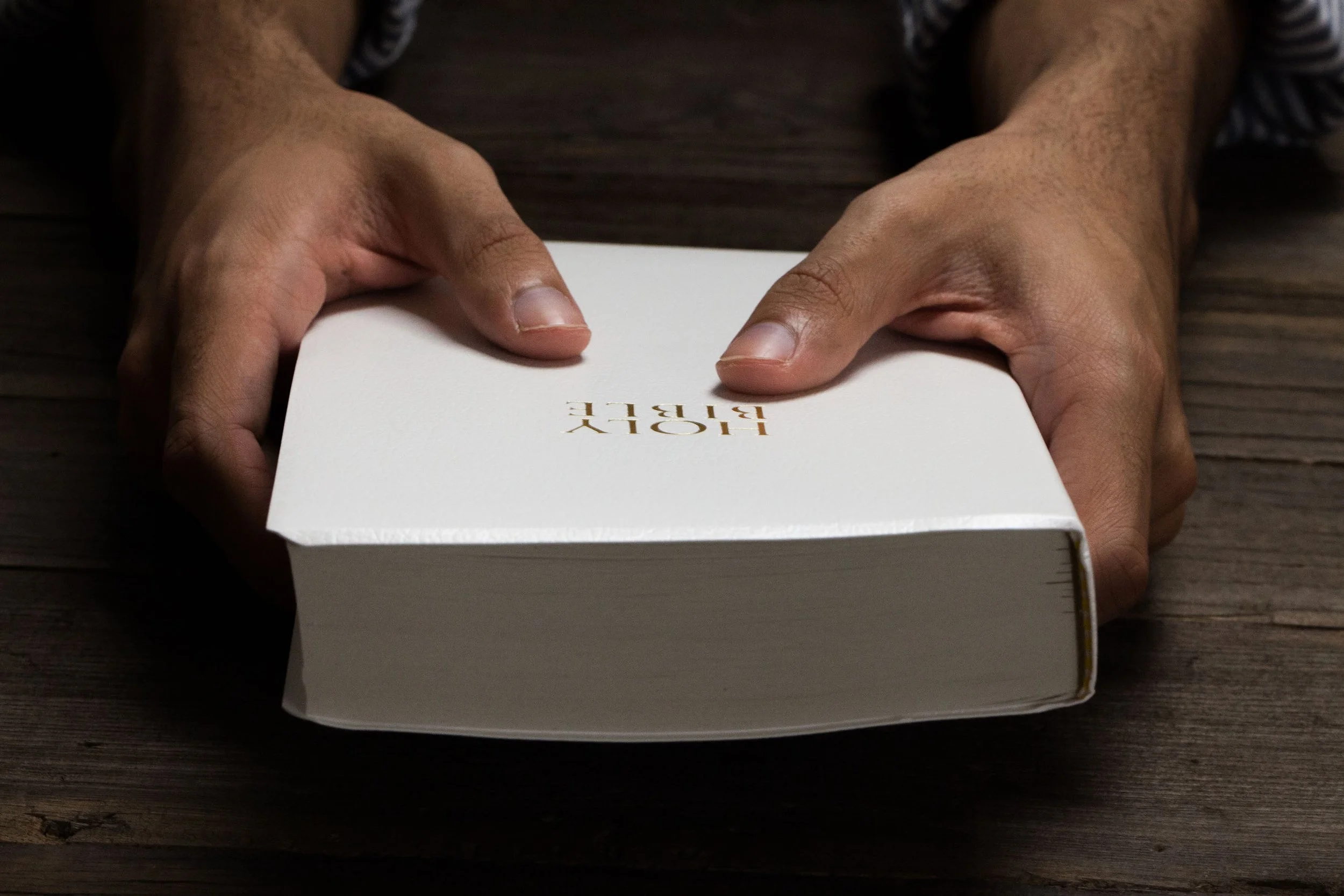 Person holding a white Bible with gold embossed text, sitting at a wooden table.