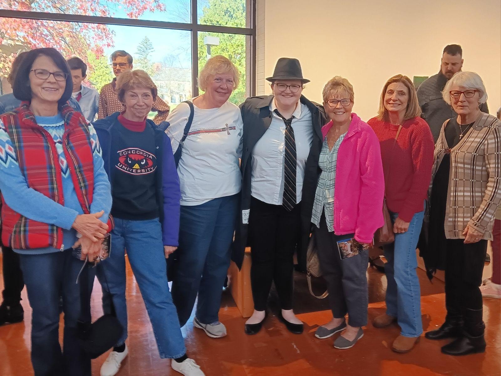 A group of women and men gathered indoors, smiling at the camera, with a large window showing trees and a clear sky behind them.