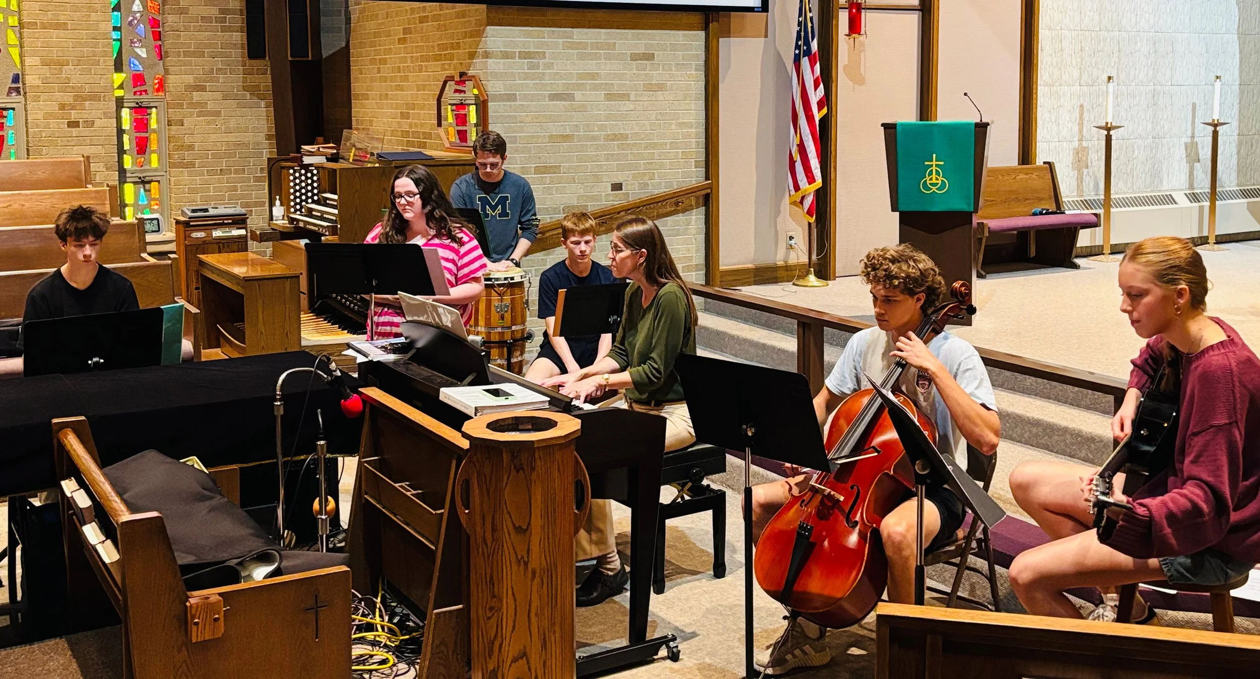 A group of young musicians rehearsing in a church, with a woman playing the keyboard, a young man playing the cello, and several other children playing musical instruments.