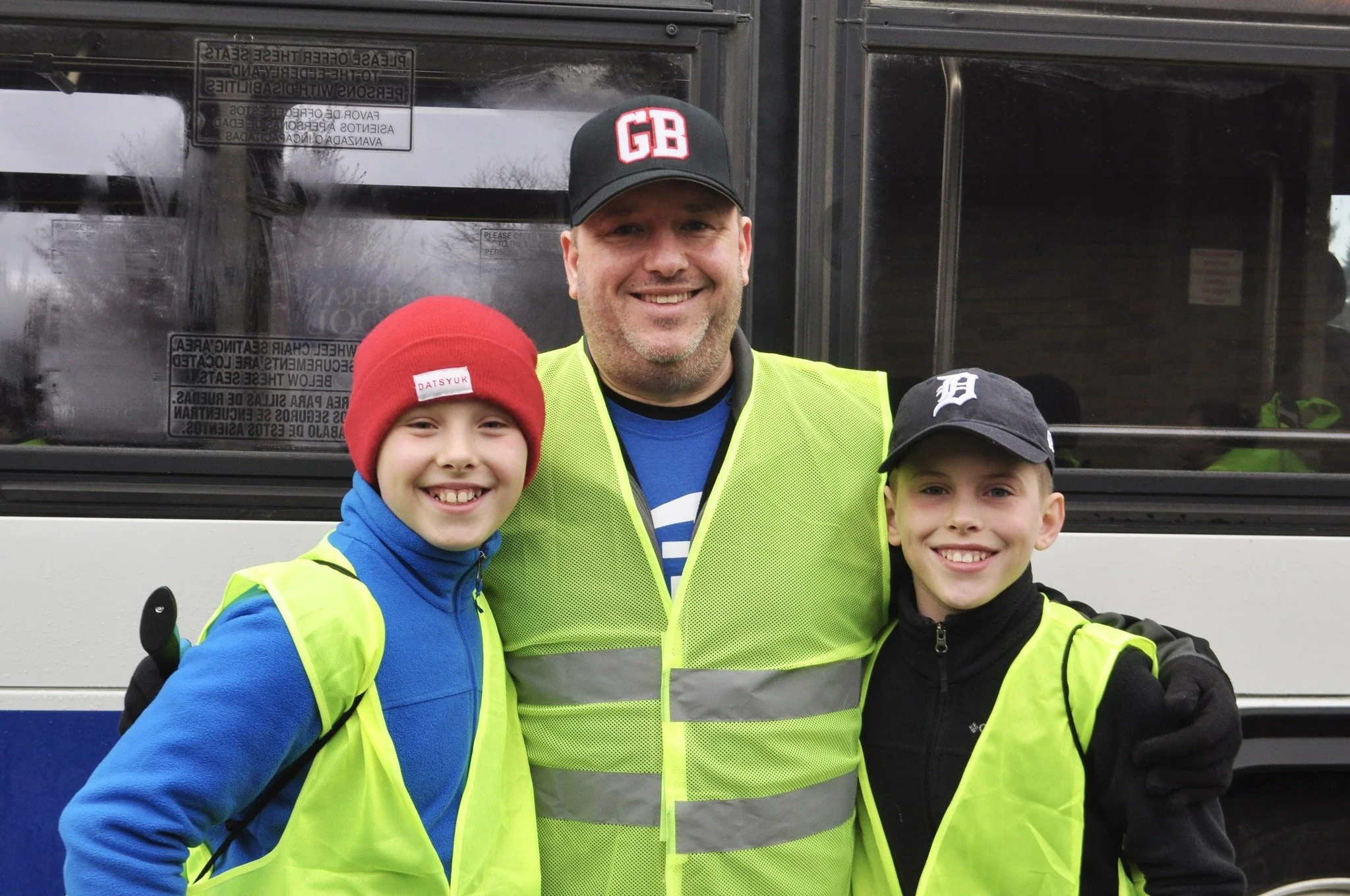 Three smiling people, an adult man and two children, wearing bright yellow safety vests standing in front of a bus, outdoors during daytime