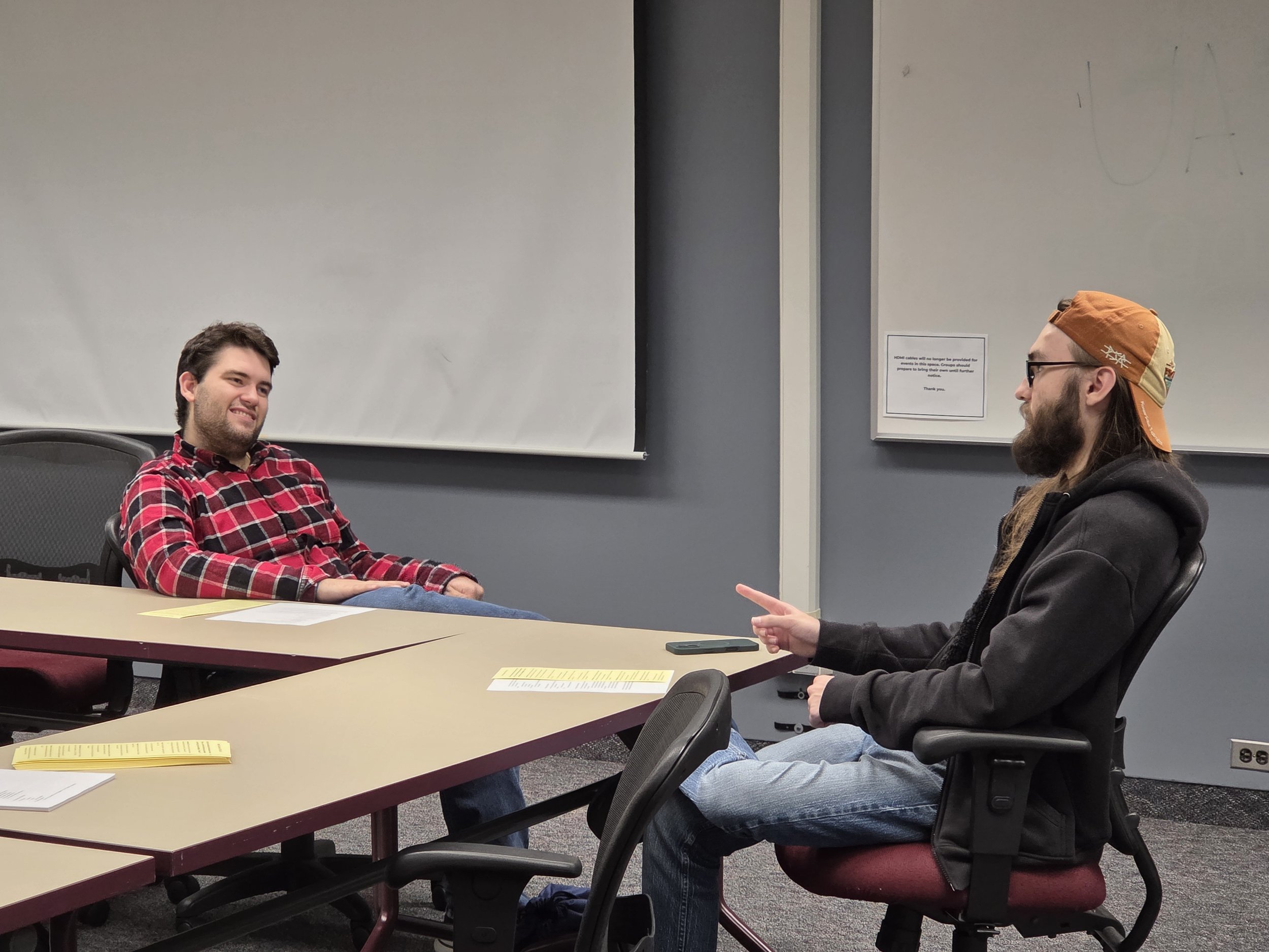 Two men having a casual conversation in a conference room, one with a red plaid shirt smiling and the other with a black hoodie and orange cap gesturing with his finger.