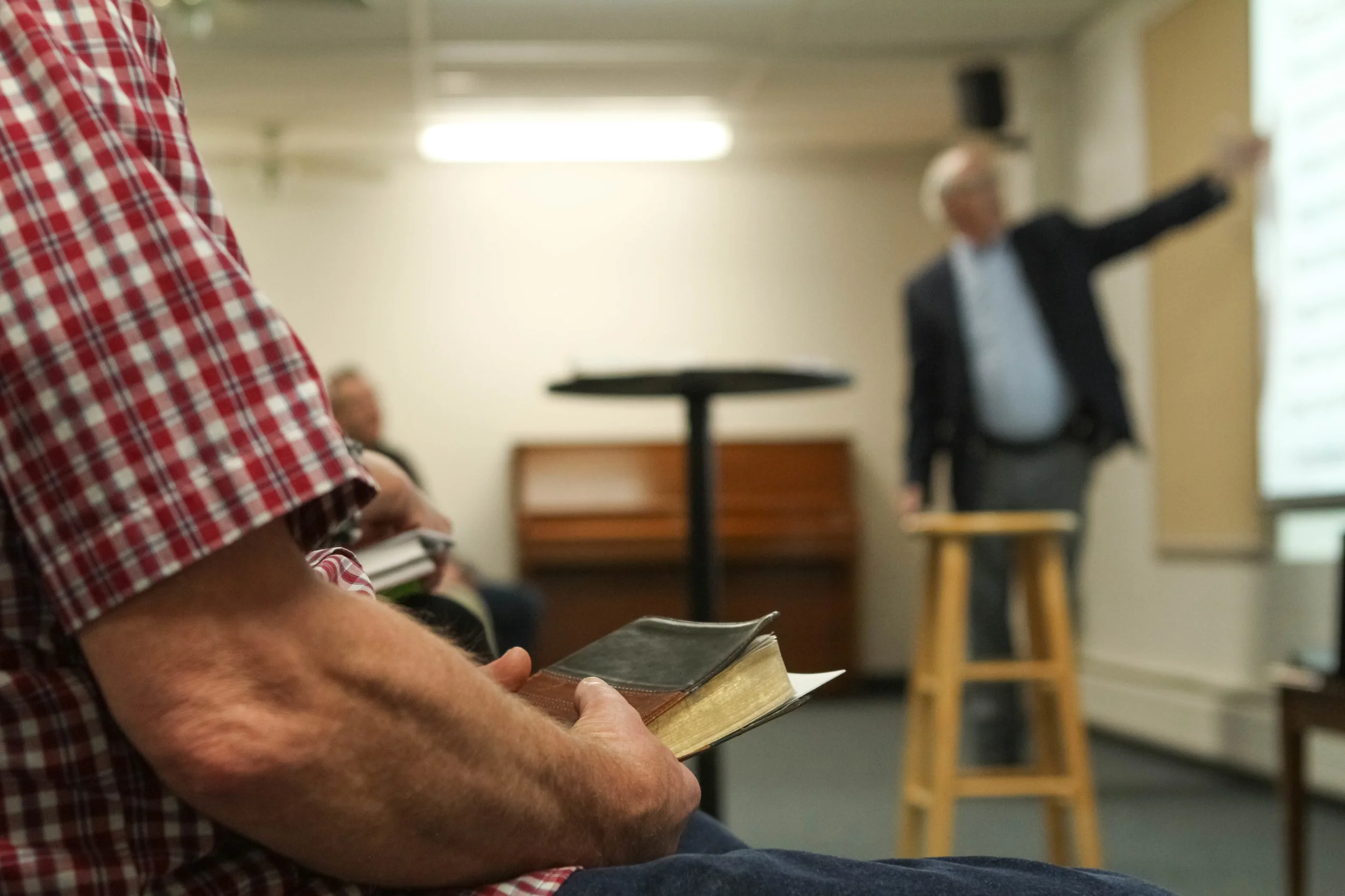 Person in red checkered shirt holding a book, seated in a classroom or seminar room, with a presenter standing near a window in the background.