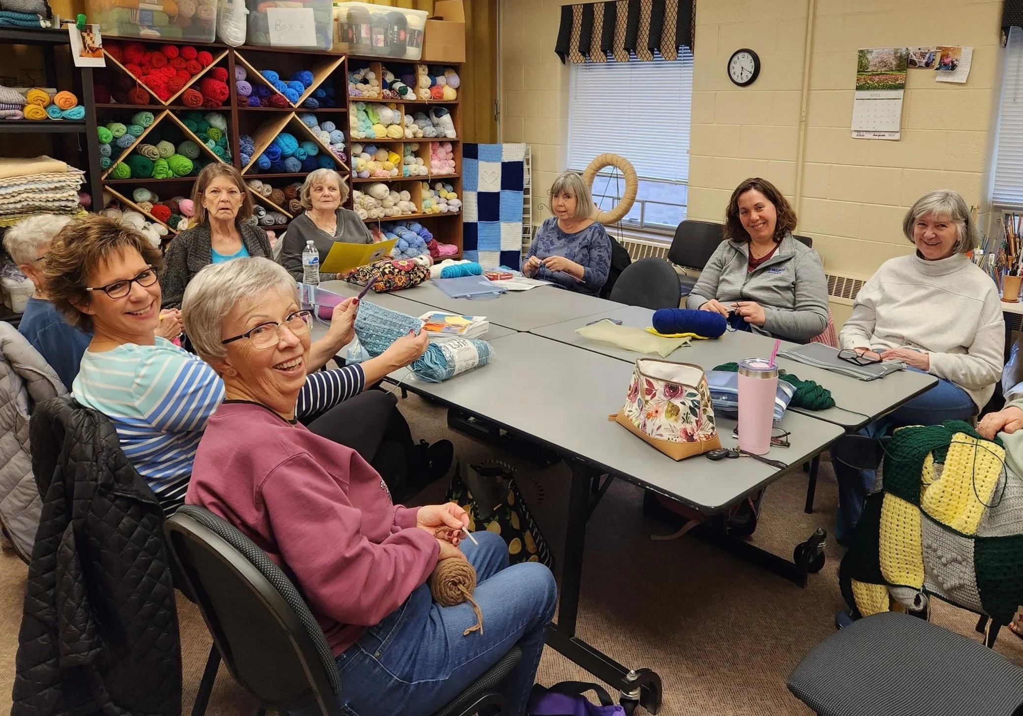 A group of women sitting around a table in a knitting or craft club room, with yarn and knitting supplies on the table and shelves full of colorful yarn in the background.
