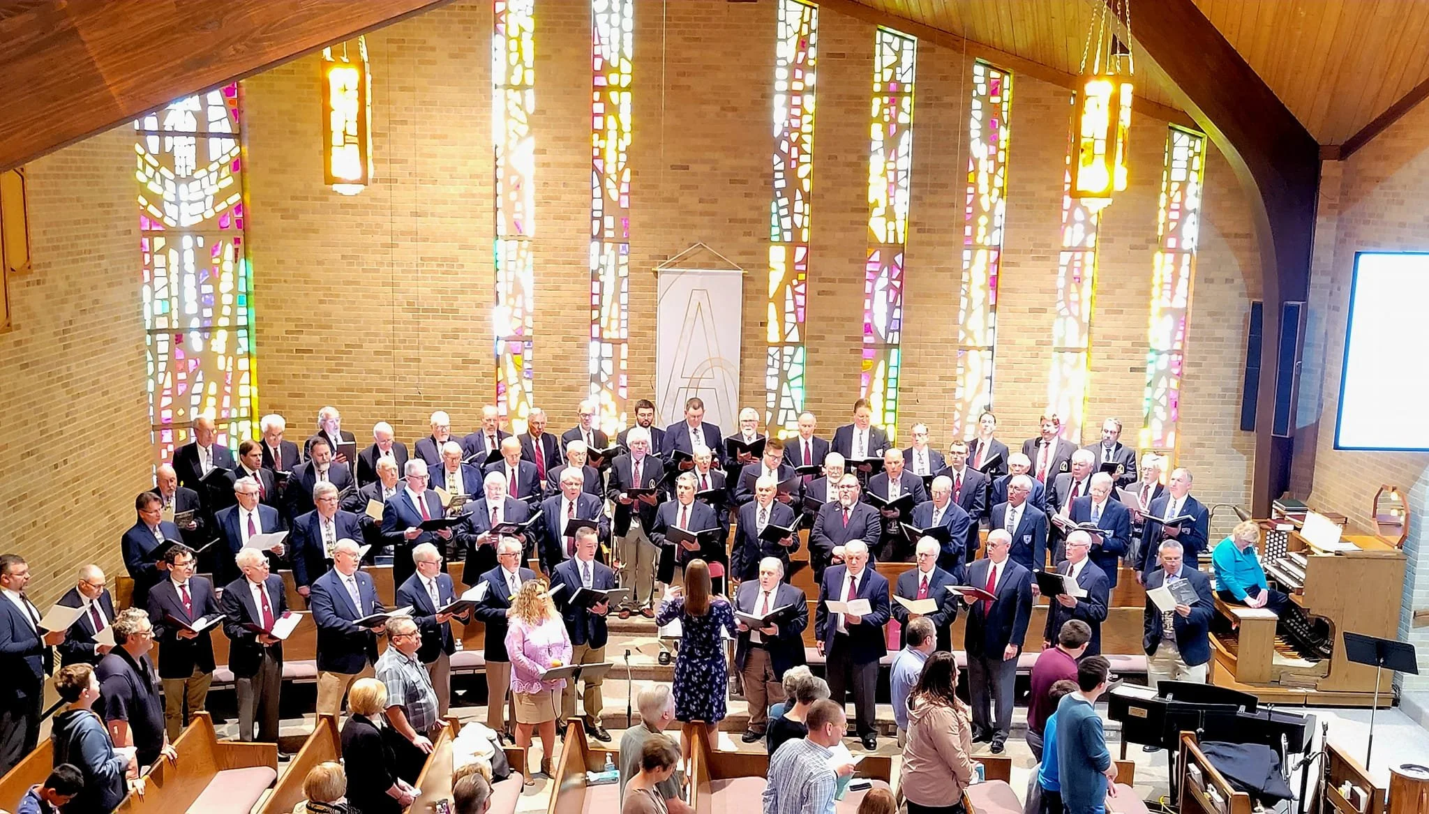 Large choir of men and women in formal suits and dresses standing on risers inside a church with stained glass windows, singing during a service or event.
