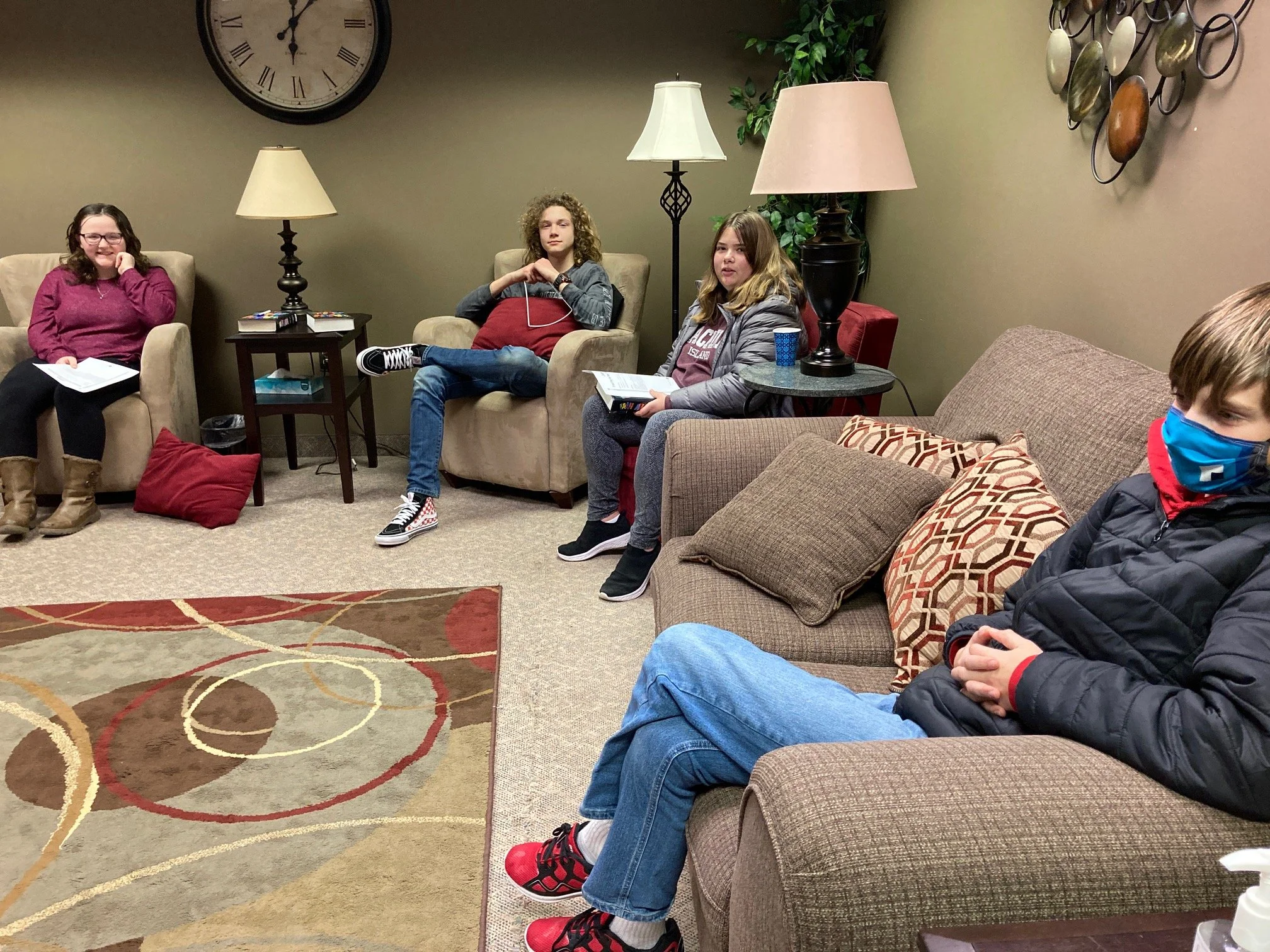 Four teenagers seated in a waiting room, three on a beige sofa and one on a beige armchair, engaging in conversation. The room has a large wall clock, two lamps, some decorative pillows, and a carpeted floor.