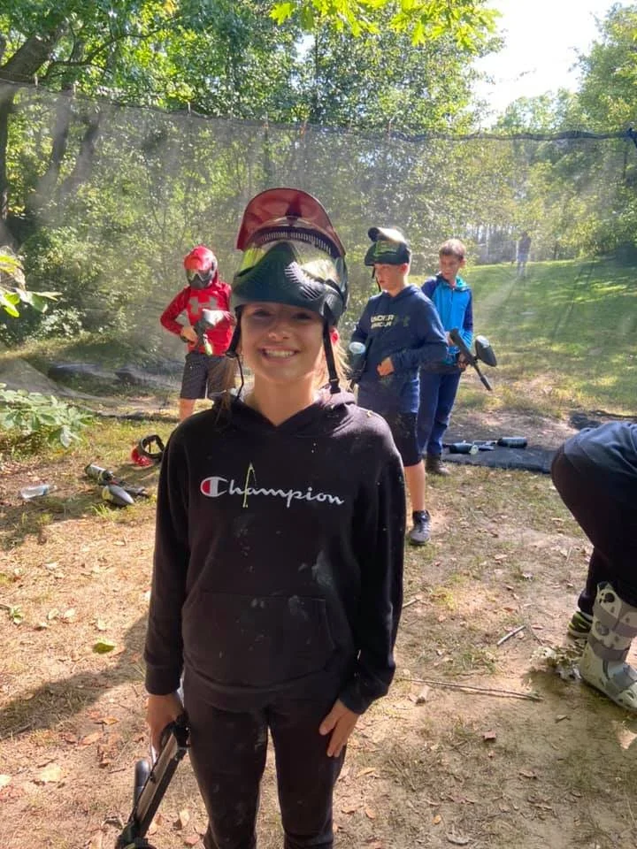 Group of children outdoors in a wooded area, some holding paintball guns and wearing helmets, with green trees and a netted barrier in the background.