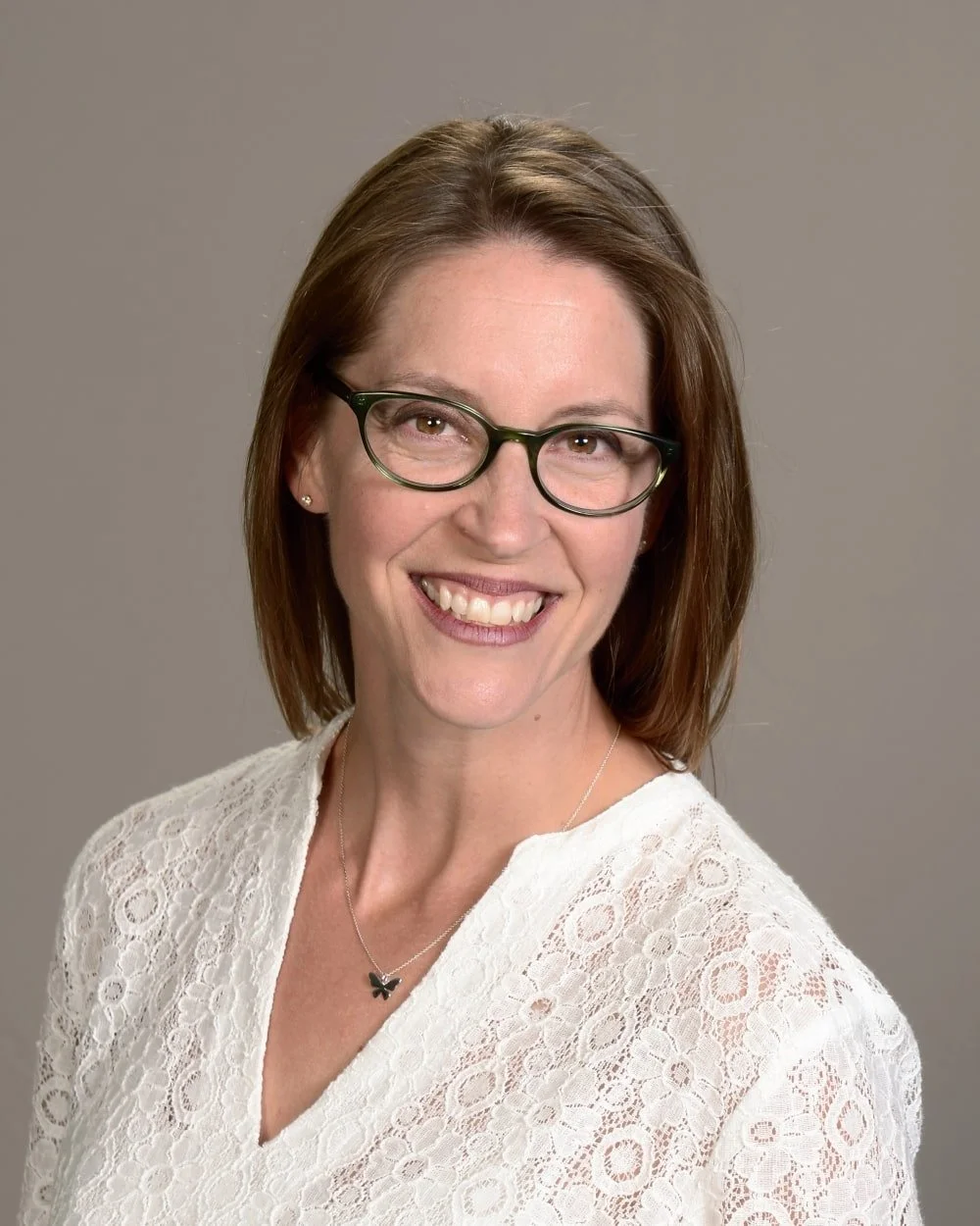 A woman with brown hair, glasses, and a white lace top, smiling at the camera against a gray background.