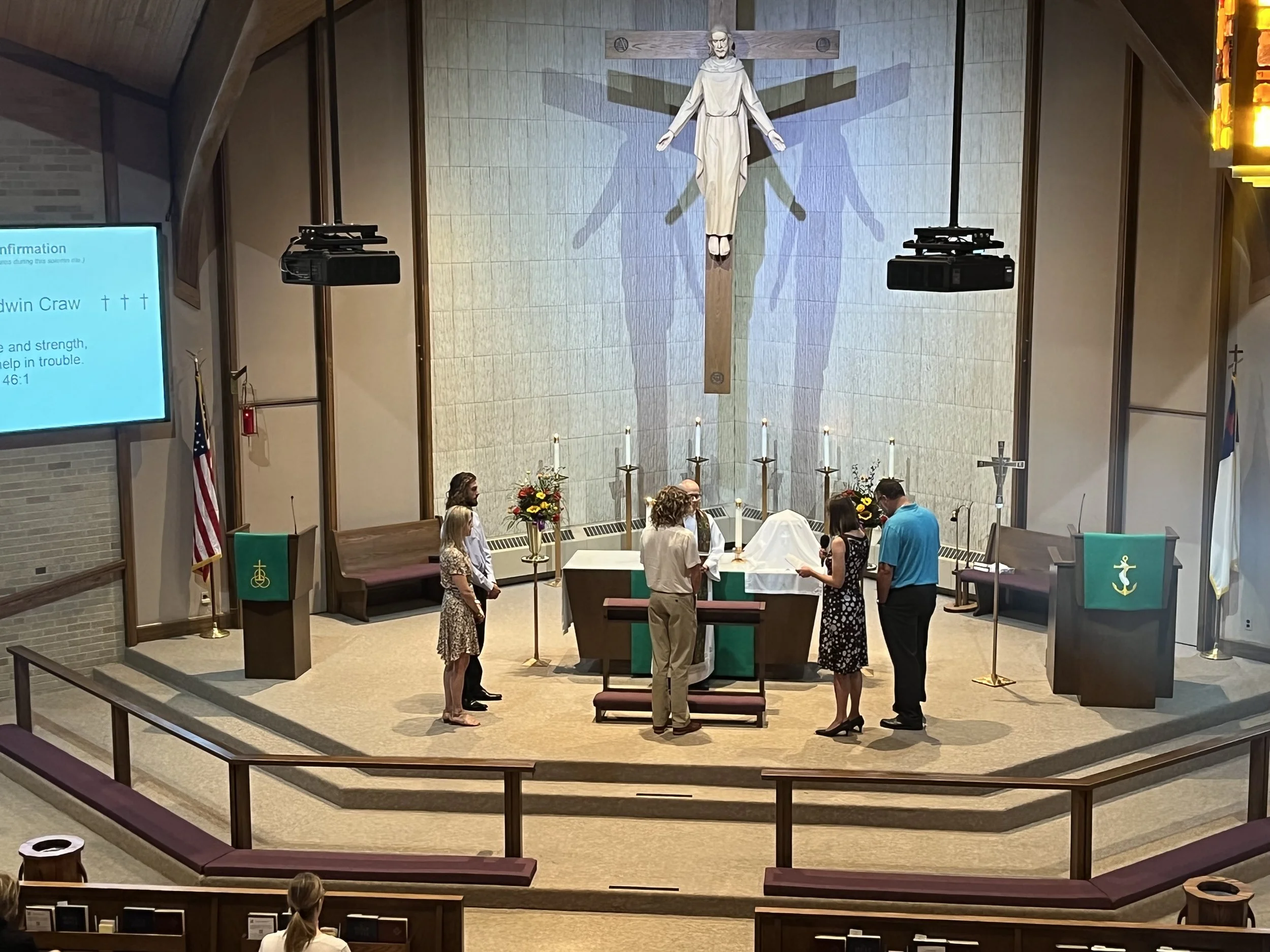 A church altar with five people gathered around it, participating in a religious ceremony during a live service in a church with a large crucifix of Jesus Christ on the wall behind.