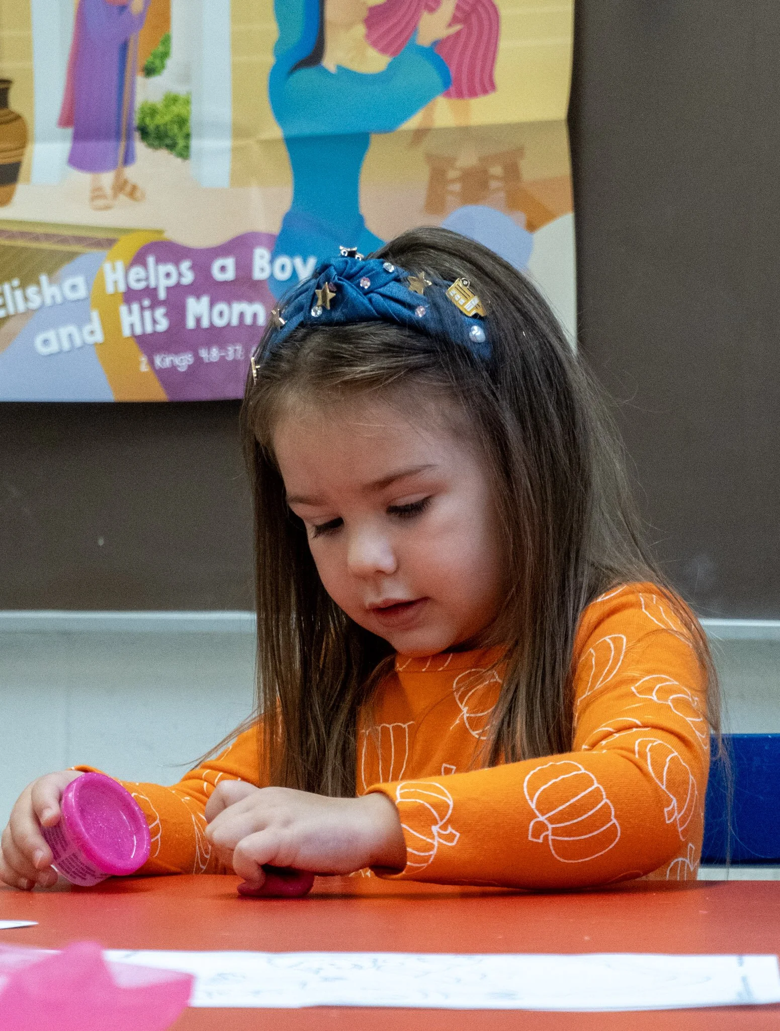 A young girl with long brown hair focused on a pink toy on a red table, wearing an orange shirt with white pumpkin patterns and a blue headband with charms, in a classroom with a colorful poster in the background.