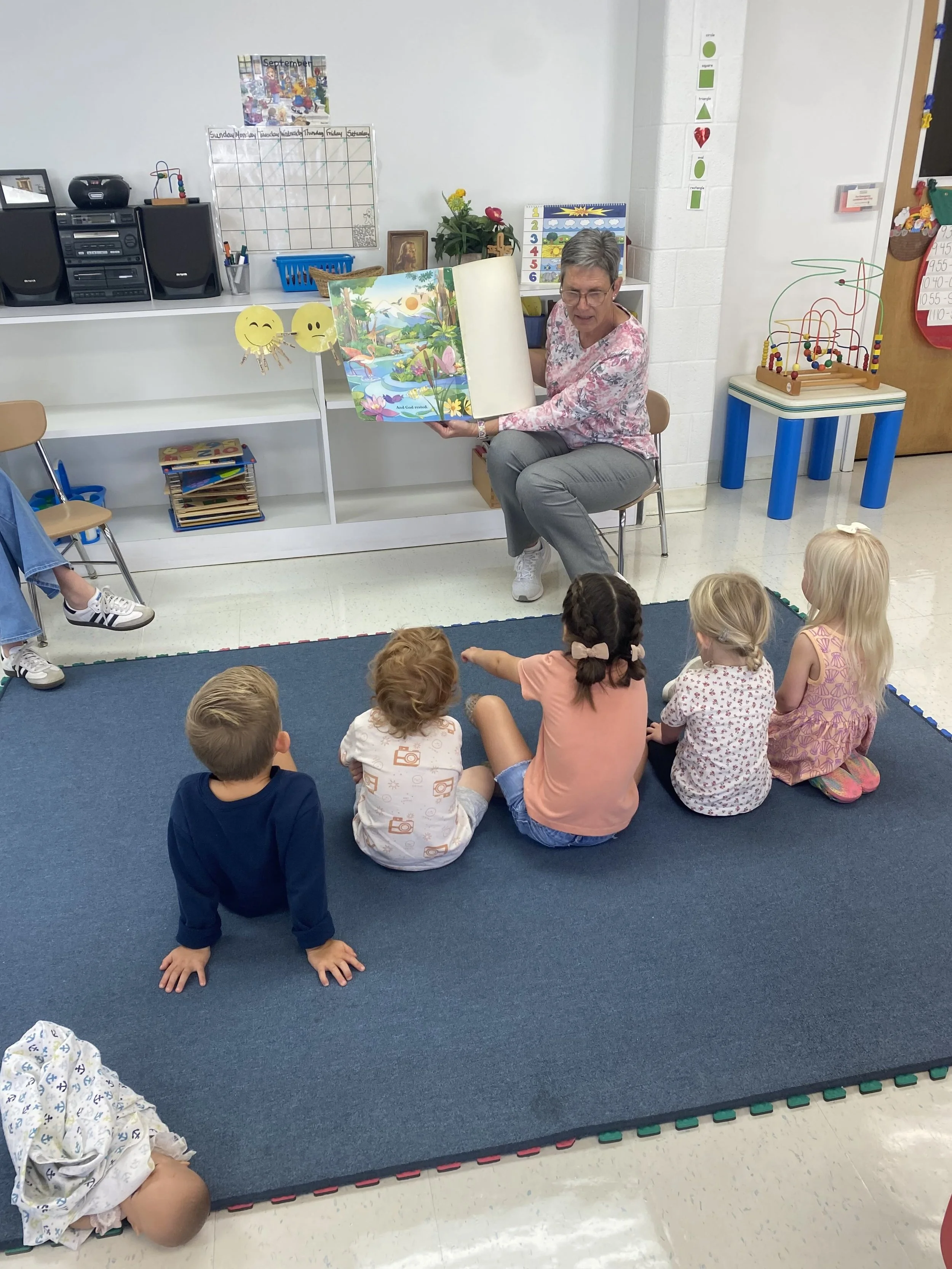 A teacher reading a colorful picture book to a group of five young children sitting on a blue carpet in a classroom.
