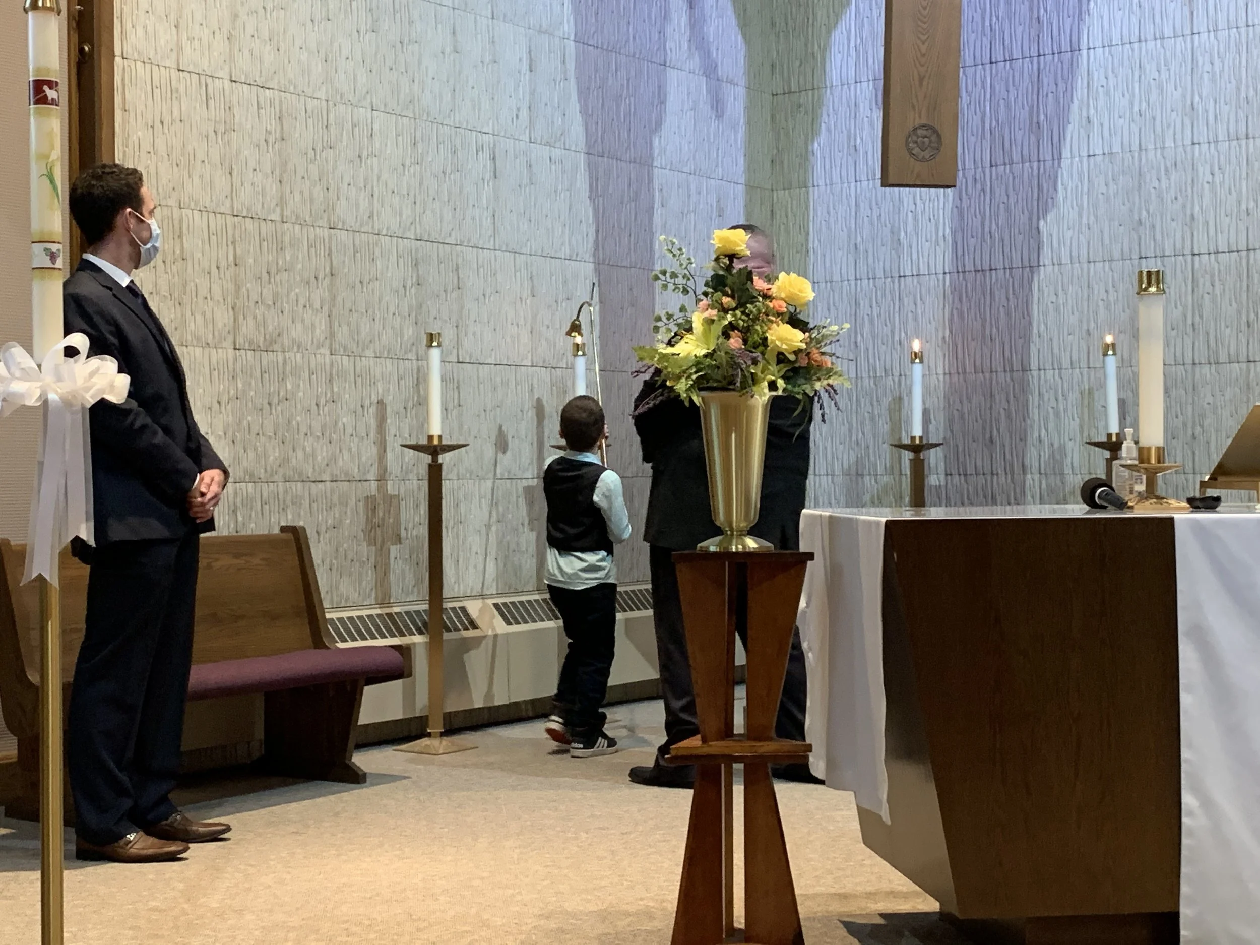 A funeral or memorial service with a man and a child standing near a casket while a priest offers a blessing. Candles are lit on the altar, and a large floral arrangement is in the center of the scene.