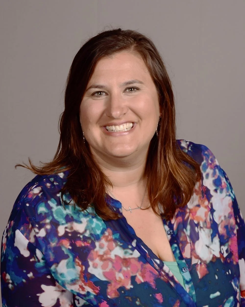 A woman with shoulder-length auburn hair smiling, wearing a colorful blue and floral blouse, against a gray background.