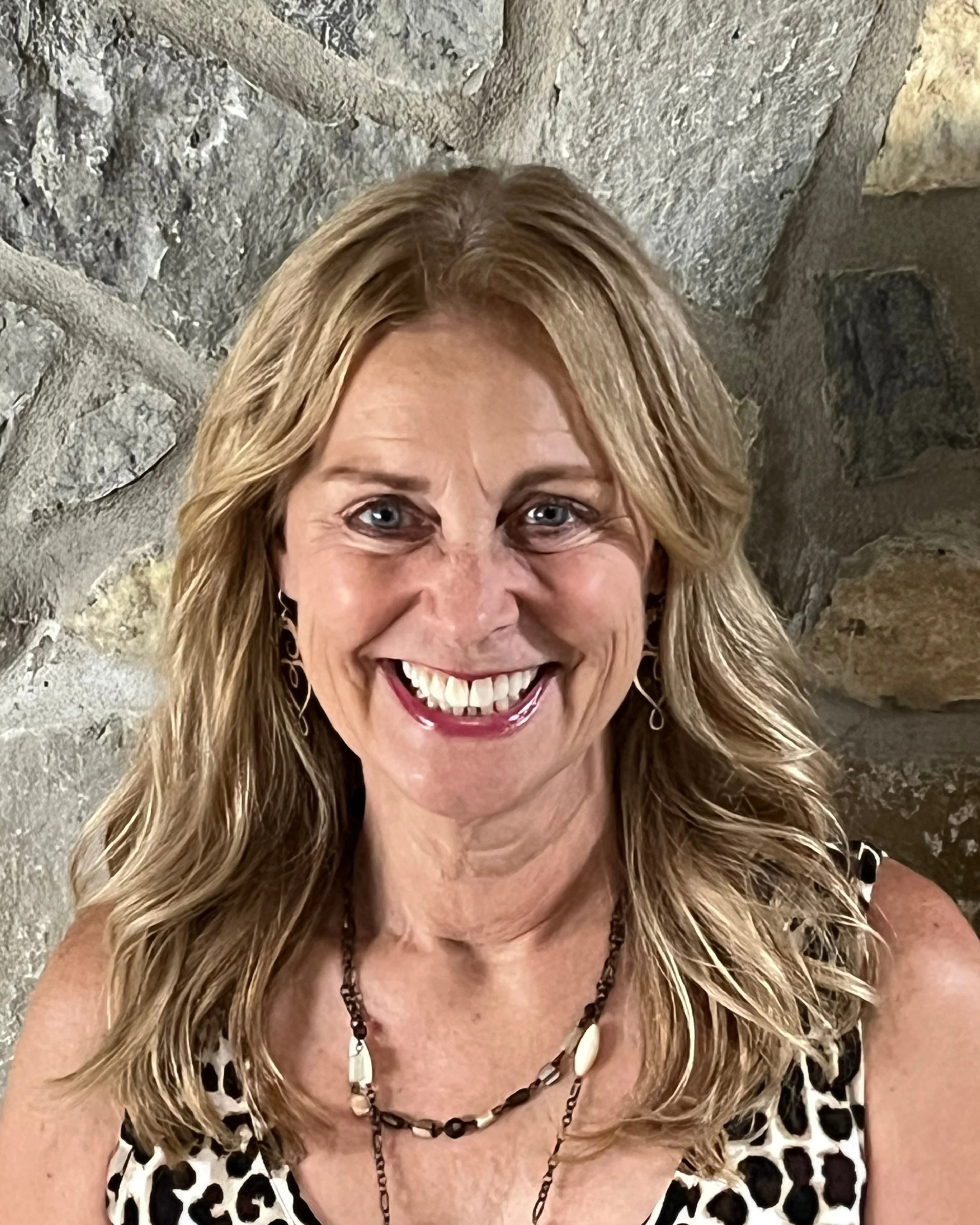 A woman with blonde hair, blue eyes, and a warm smile, wearing a black top with lace-up front and silver earrings, standing against a neutral background.