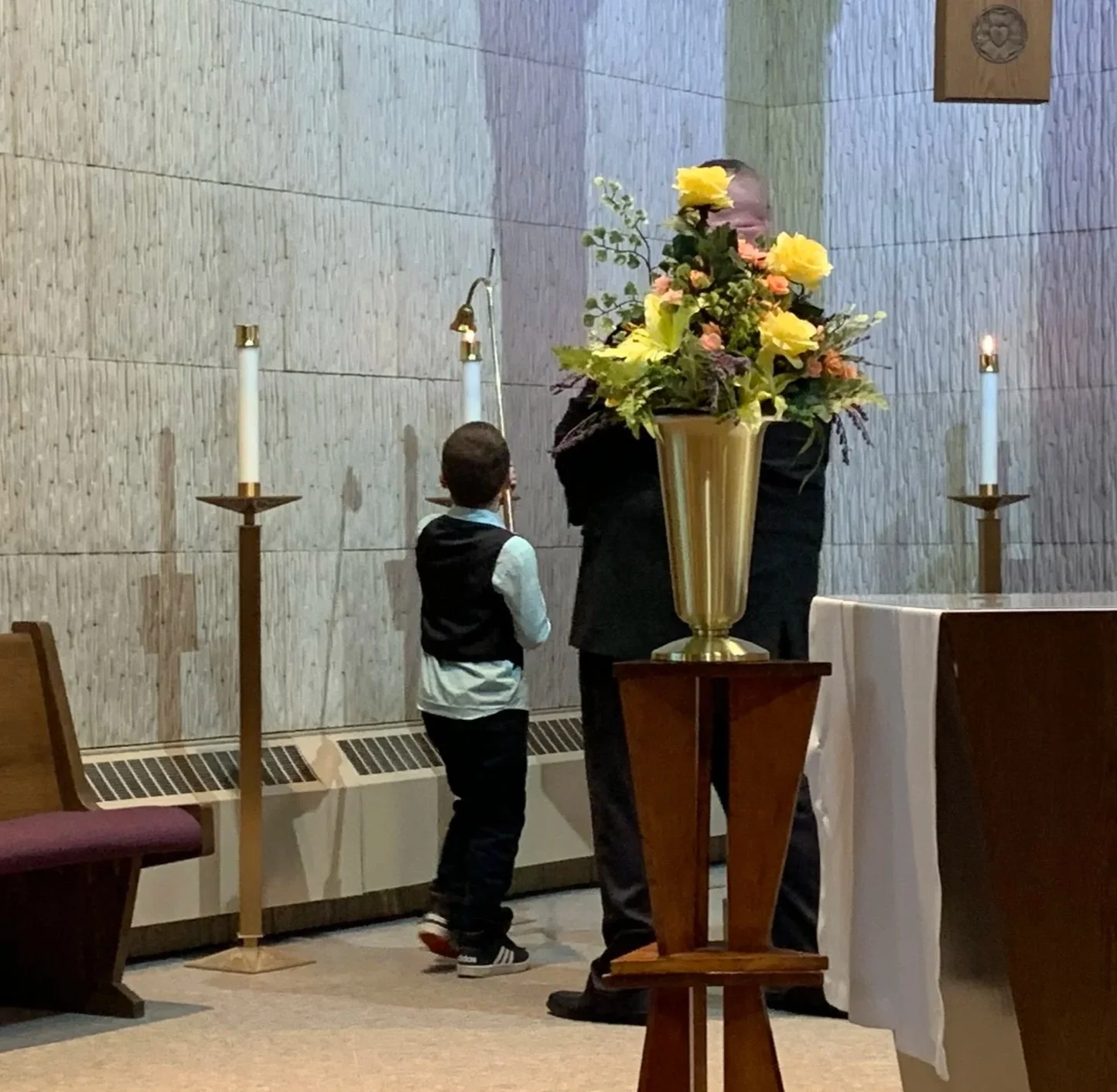 A boy and an adult standing in a chapel near an altar with a large floral arrangement and lit candles.