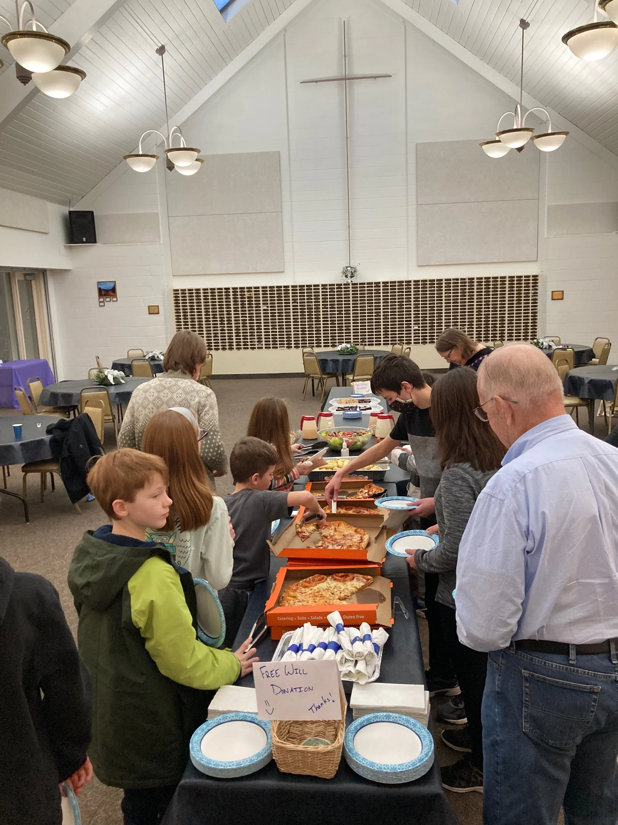 People at a pizza buffet in a church hall with a cross on the wall, children and adults lining up to serve and eat pizza, and a sign indicating free donation for the food.