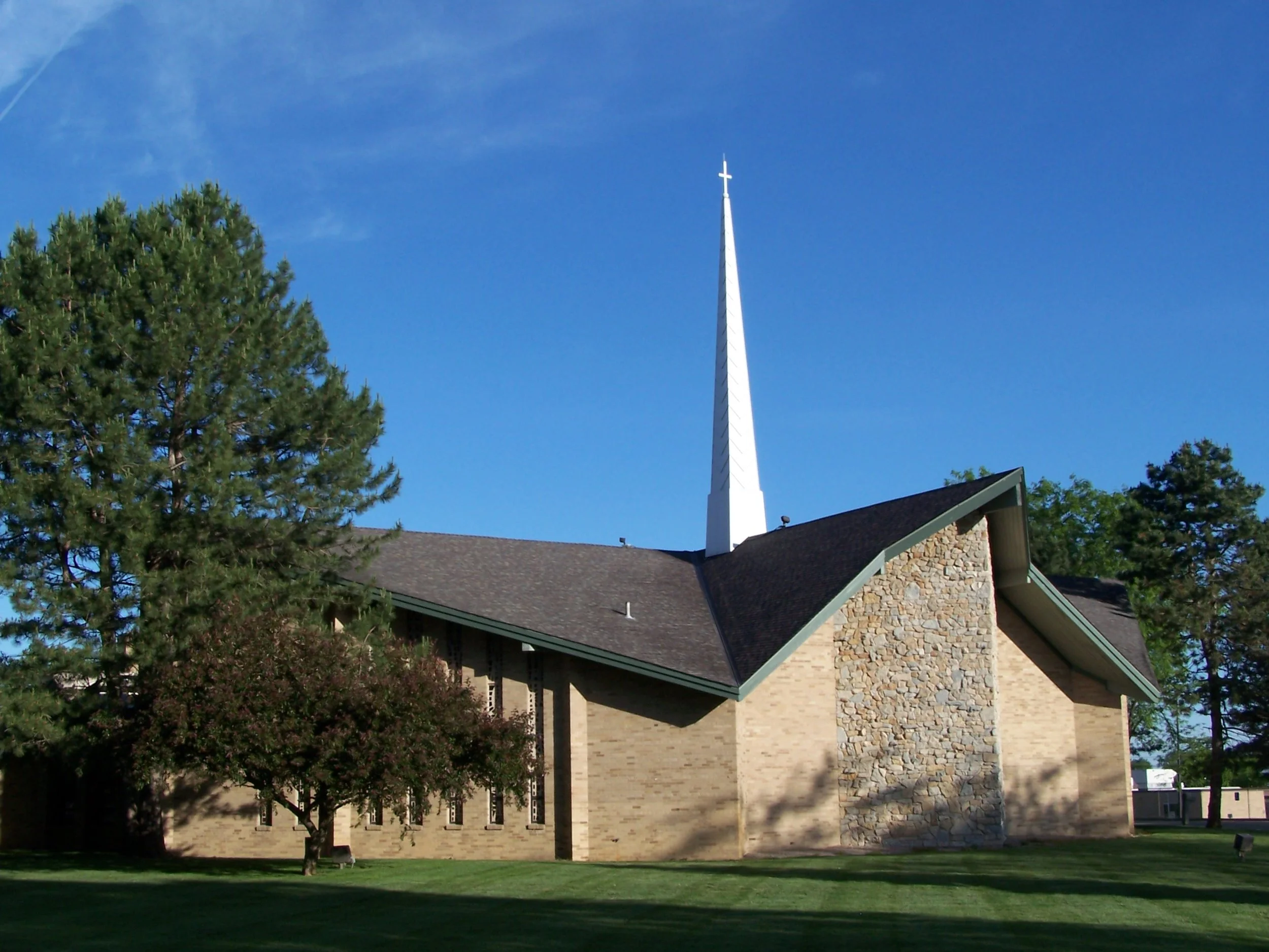 A church with a tall white steeple, beige brick walls, and a gabled roof, surrounded by trees and a grassy lawn under a blue sky.