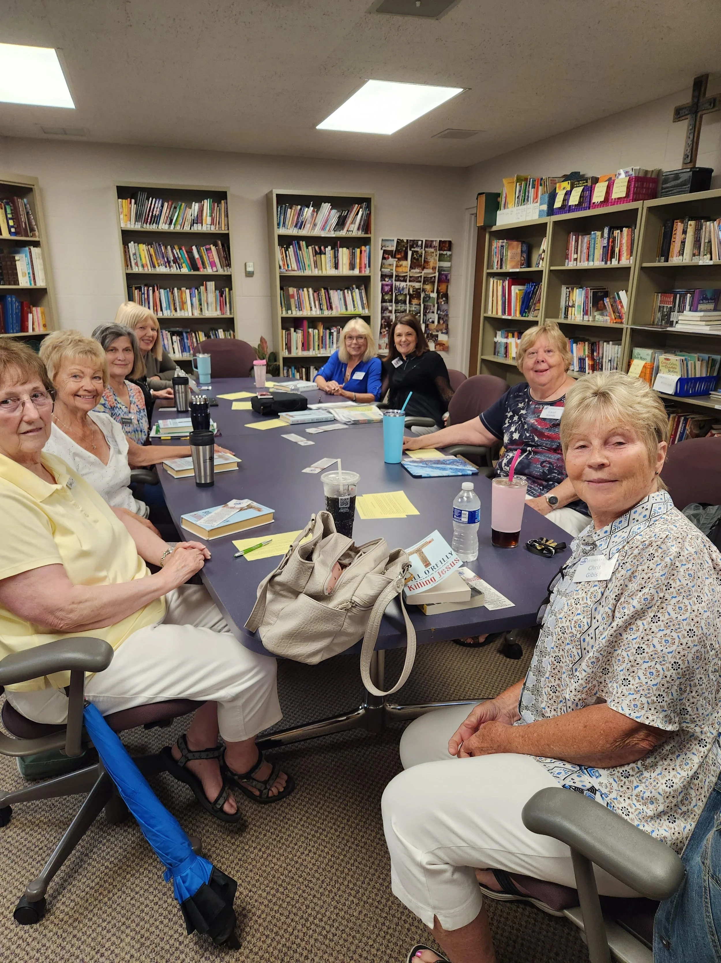 A group of women sitting around a large conference table in a room with bookshelves, engaging in a meeting or discussion.