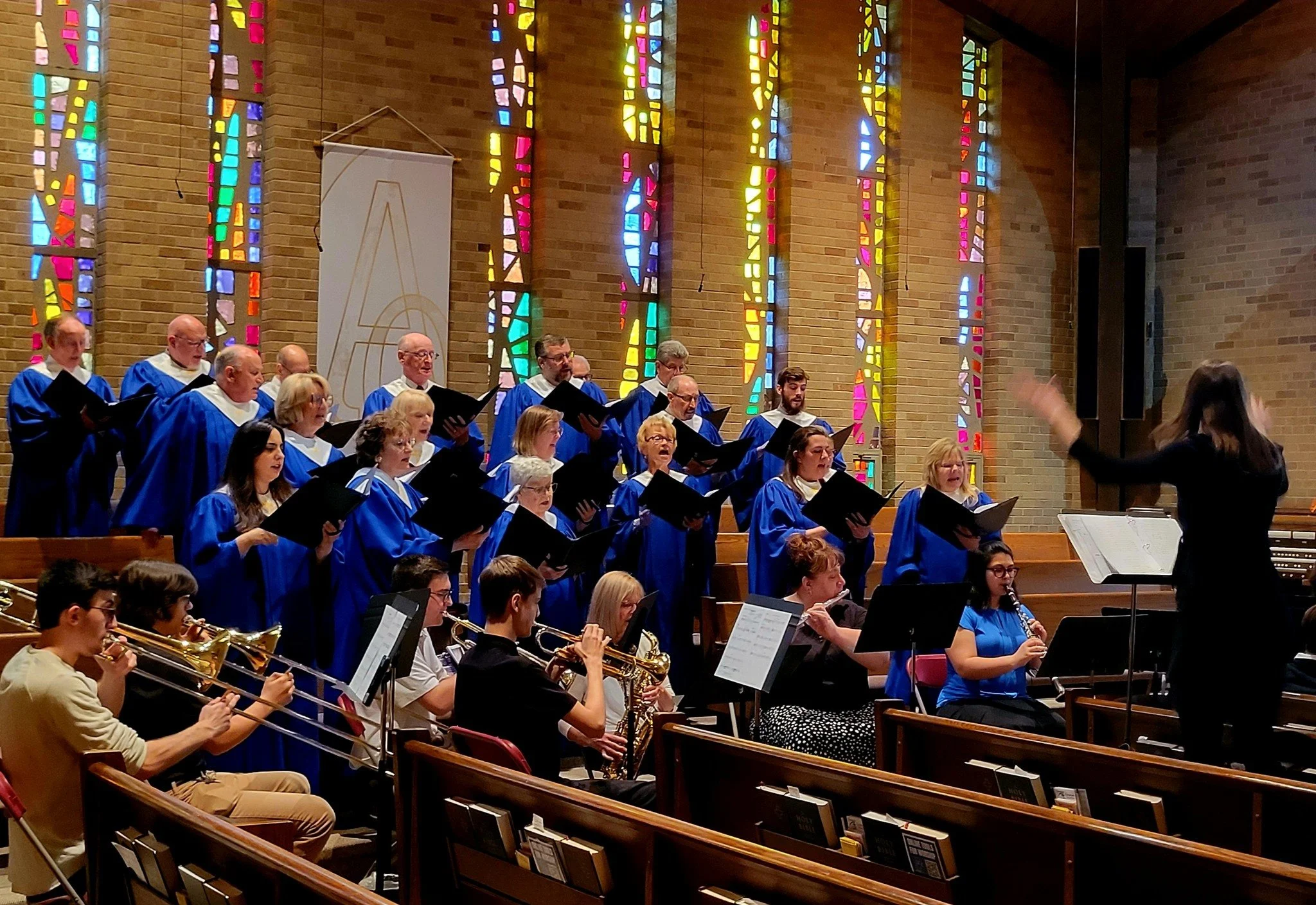 A choir and an orchestra performing inside a church with stained glass windows in the background.