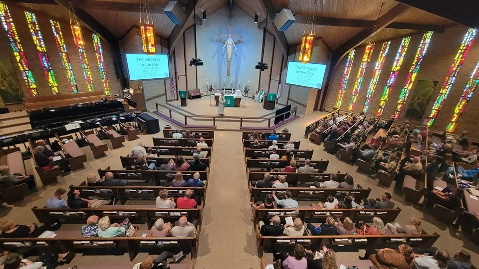 Inside a church with stained glass windows, a congregation seated in wooden pews, a cross and a statue at the altar, and screens displaying 'The Message for the Day'.