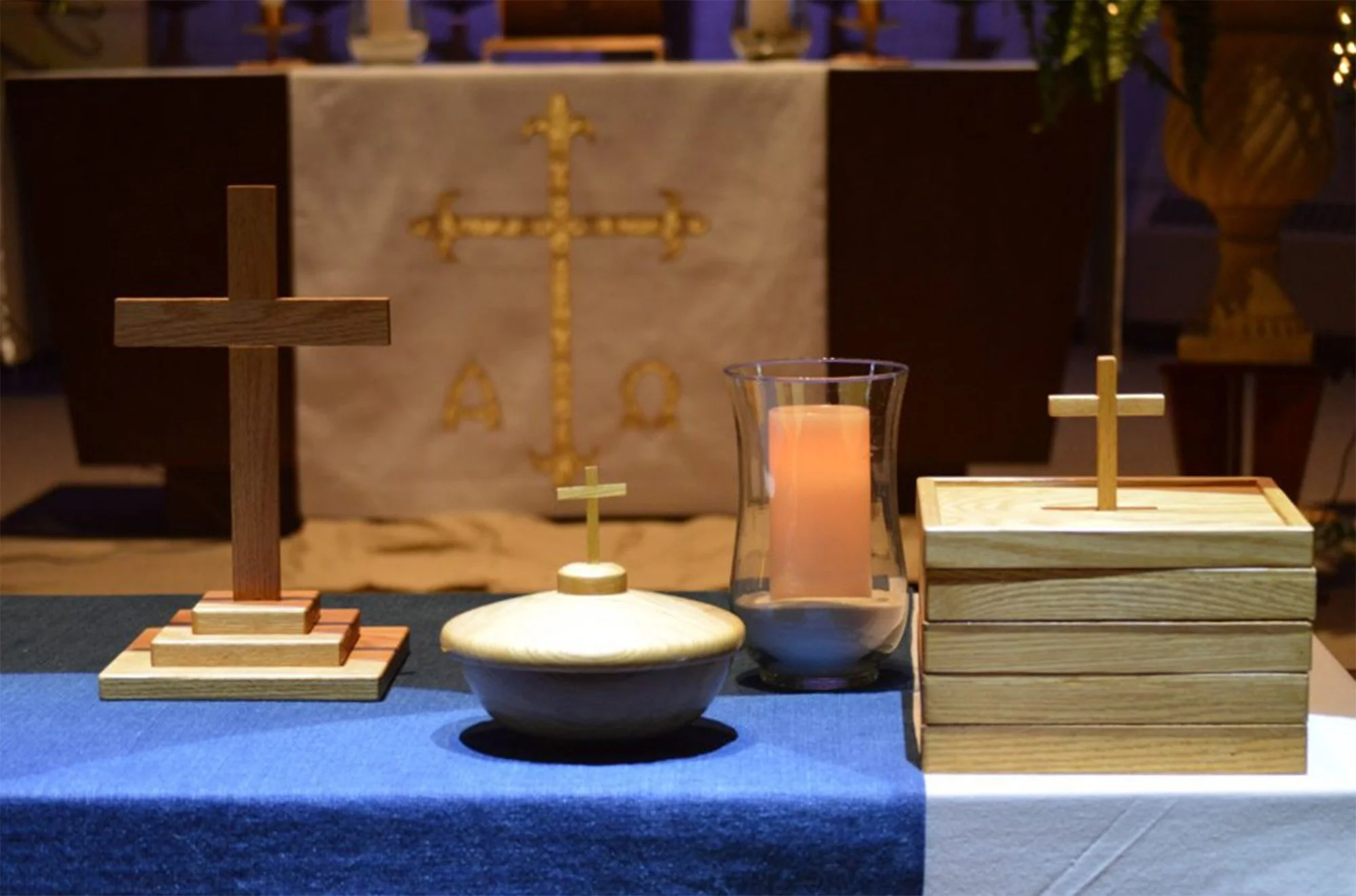 Religious altar with wooden crosses, a candle, and a cloth, featuring a backdrop with a golden cross and Greek letters, surrounded by decorative items.