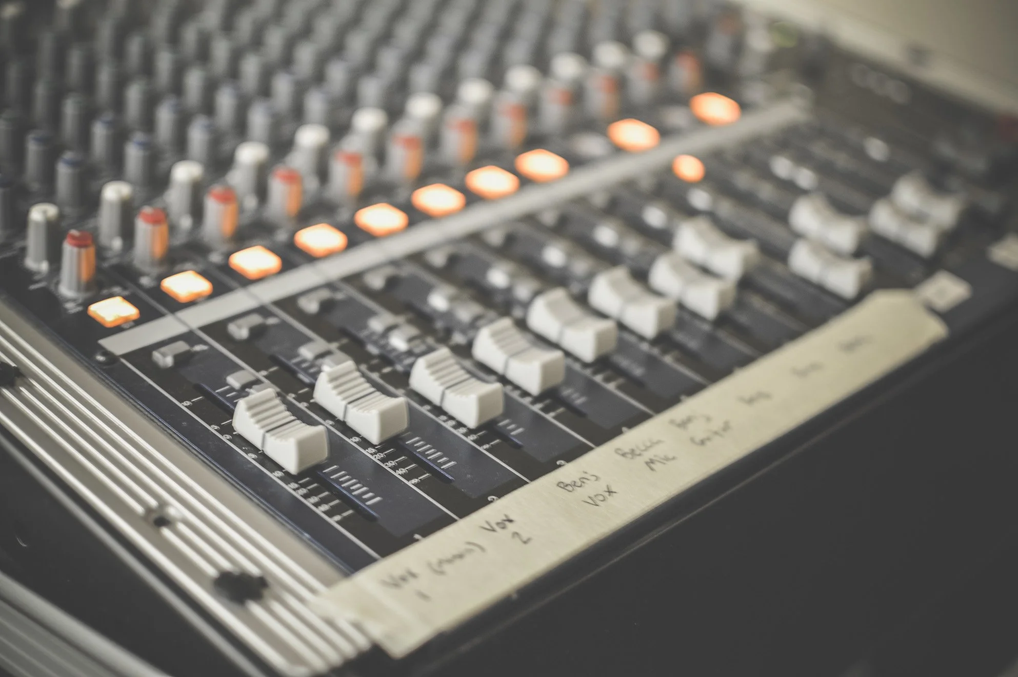 Close-up of a sound mixing console with sliders, knobs, and illuminated buttons, with handwritten labels on masking tape along the bottom edge.