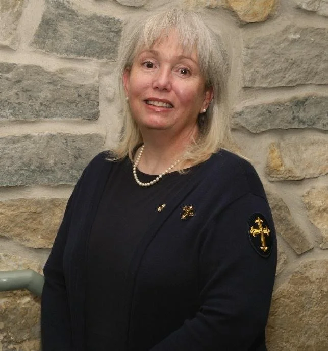 A portrait of a woman with blonde hair, wearing a blue top, a pearl necklace, and pearl earrings, smiling softly against a plain gray background.