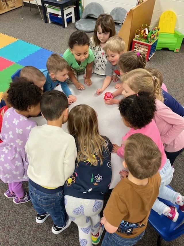 Group of young children gathered around a white table in a classroom, playing with a pink object.