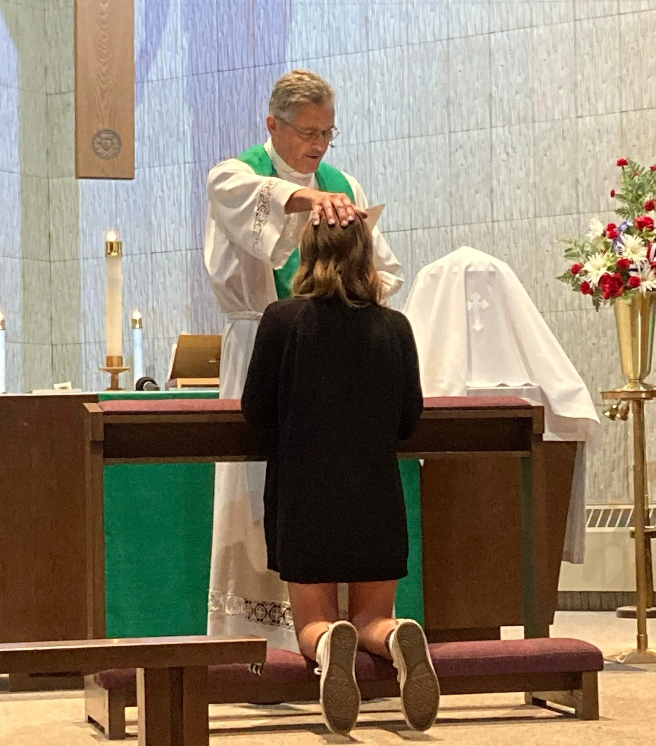 A woman kneeling at an altar during a religious ceremony, with a priest touching her head and a covered object on the altar.