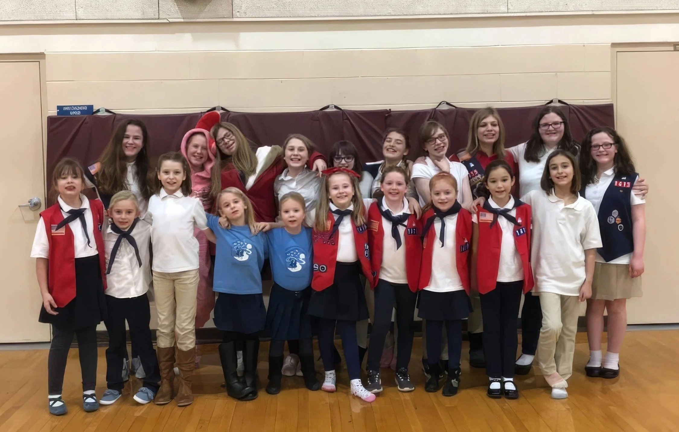 Group of children and young women posing for a photo indoors, some in Girl Scout uniforms, with a maroon curtain in the background.