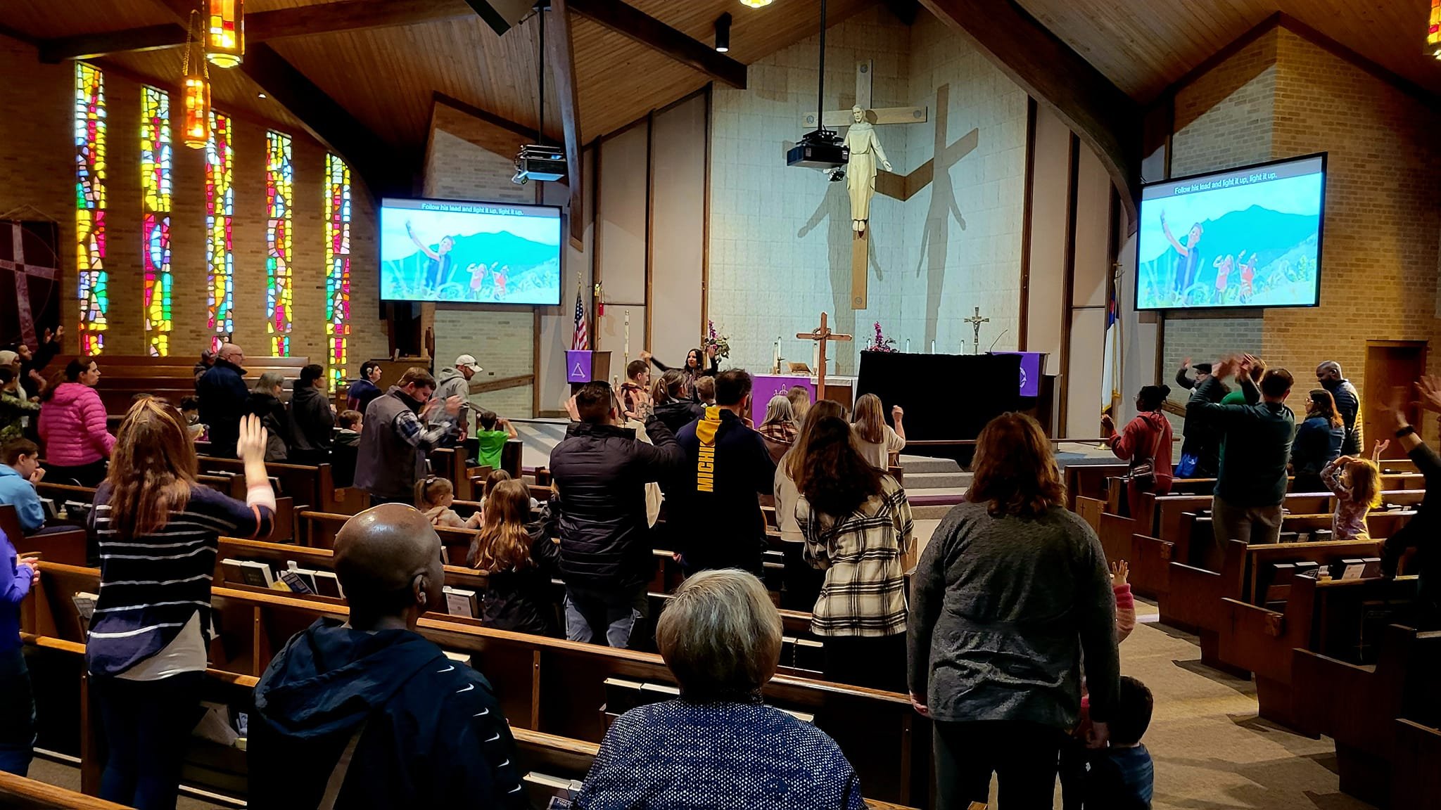People gathered in a church for a service or prayer, with some standing, raising hands, and others seated. The church features stained glass windows, large screens displaying an image and text, a wooden altar with a purple cloth, and a large insignia