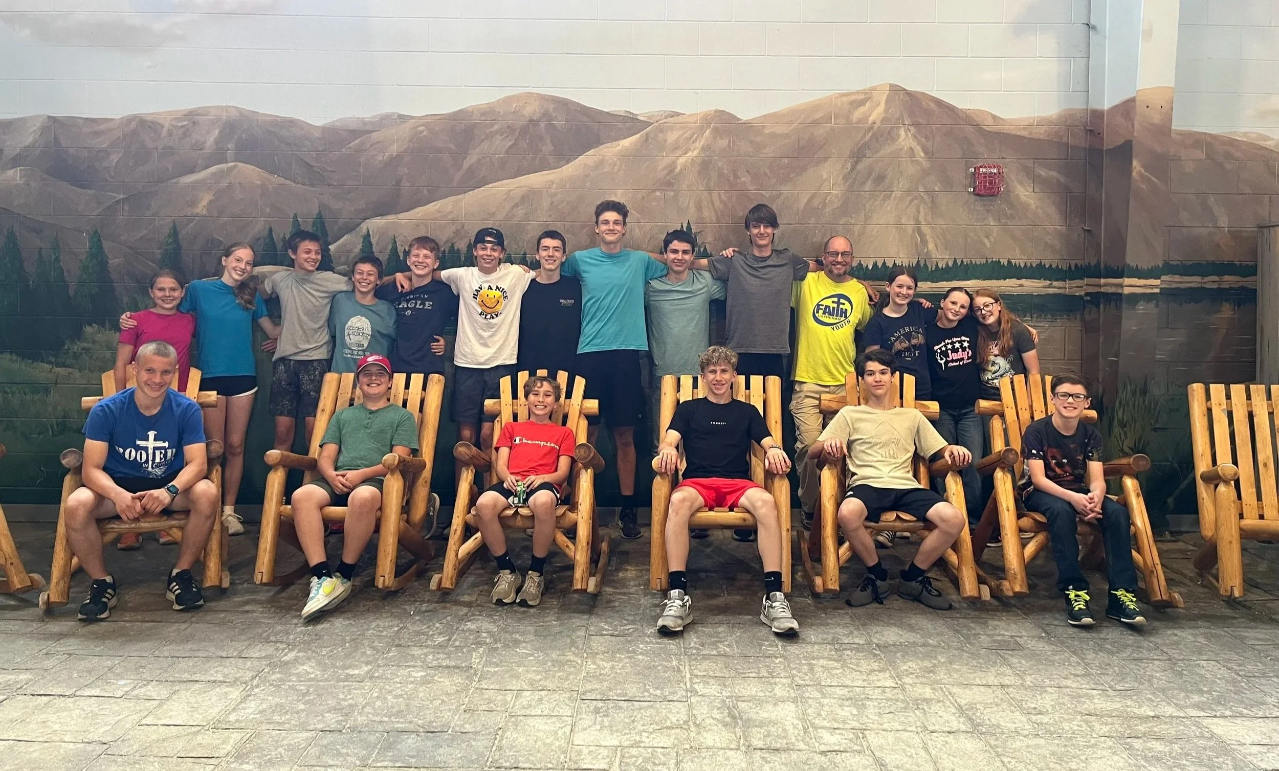 Group of children and adults smiling and posing for a photo indoors, with a mural of mountains and trees in the background, some sitting on wooden chairs and others standing behind.