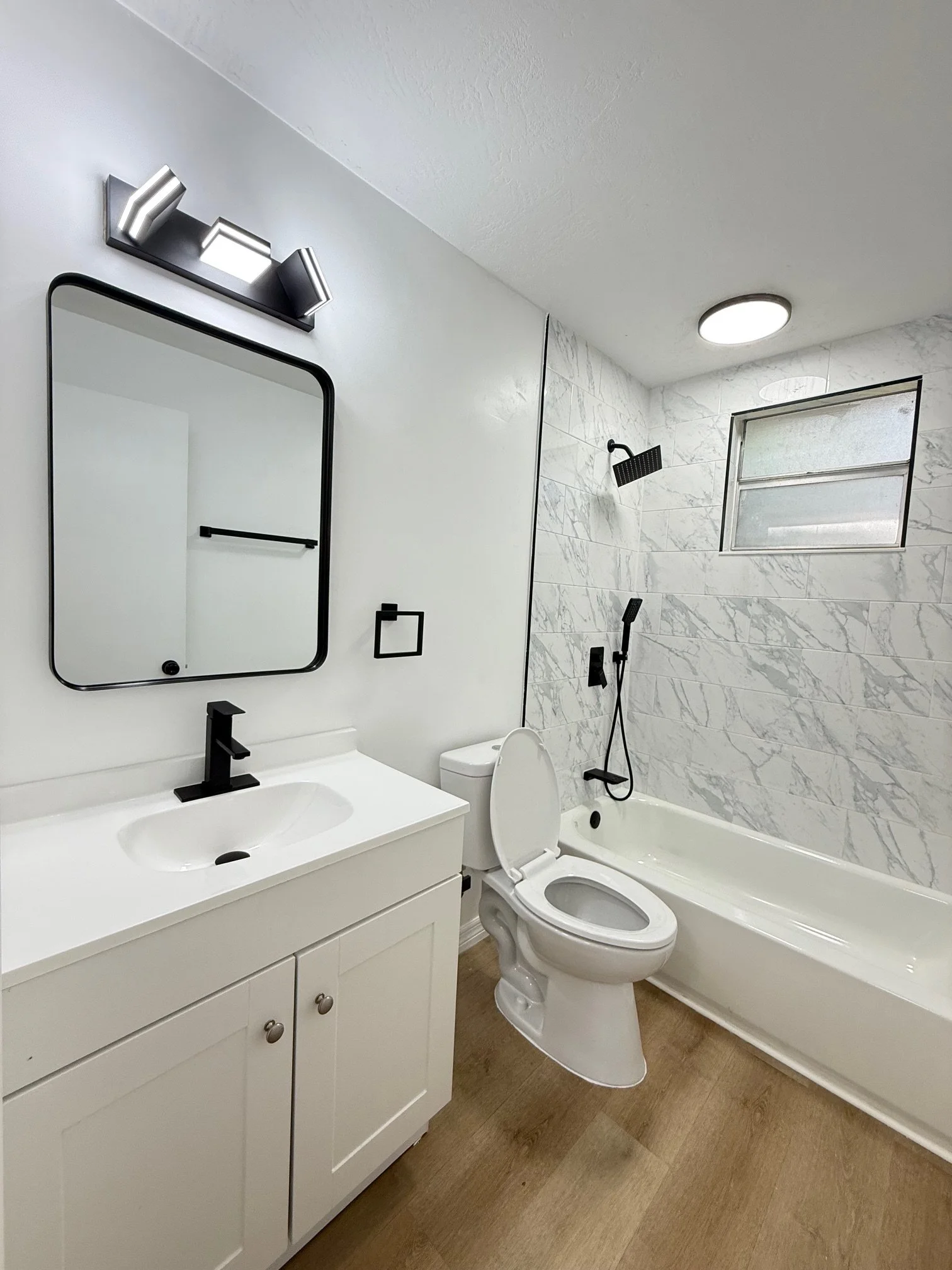 Modern bathroom with a glass-enclosed shower, white walls, a vanity with a granite countertop and blue cabinets, a large mirror, a vase with dried flowers, and shelves with small plants and decor.