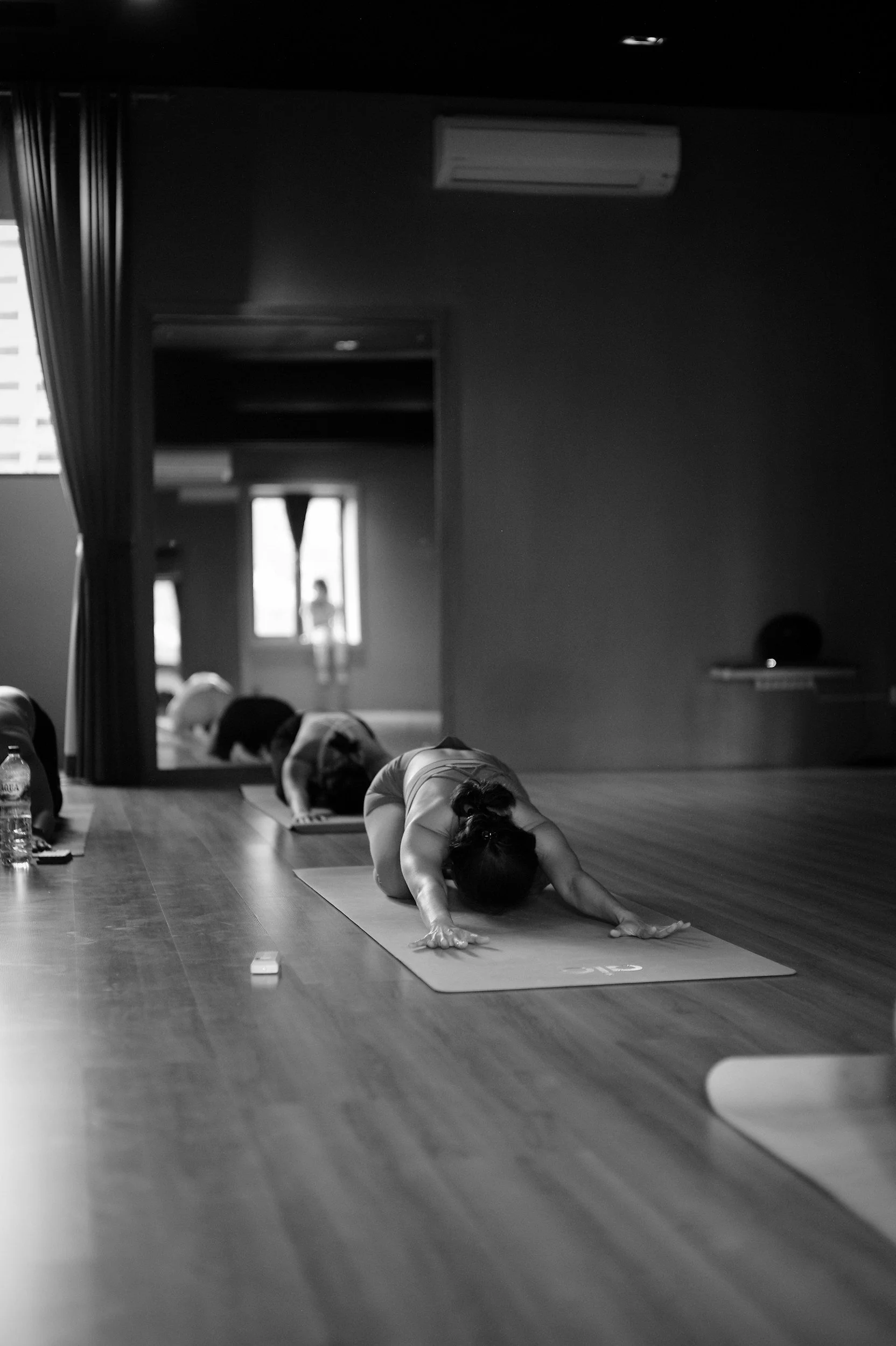 People practicing yoga in a studio, with one person in the foreground performing a forward bend pose on a yoga mat, others in the background also engaged in yoga poses.