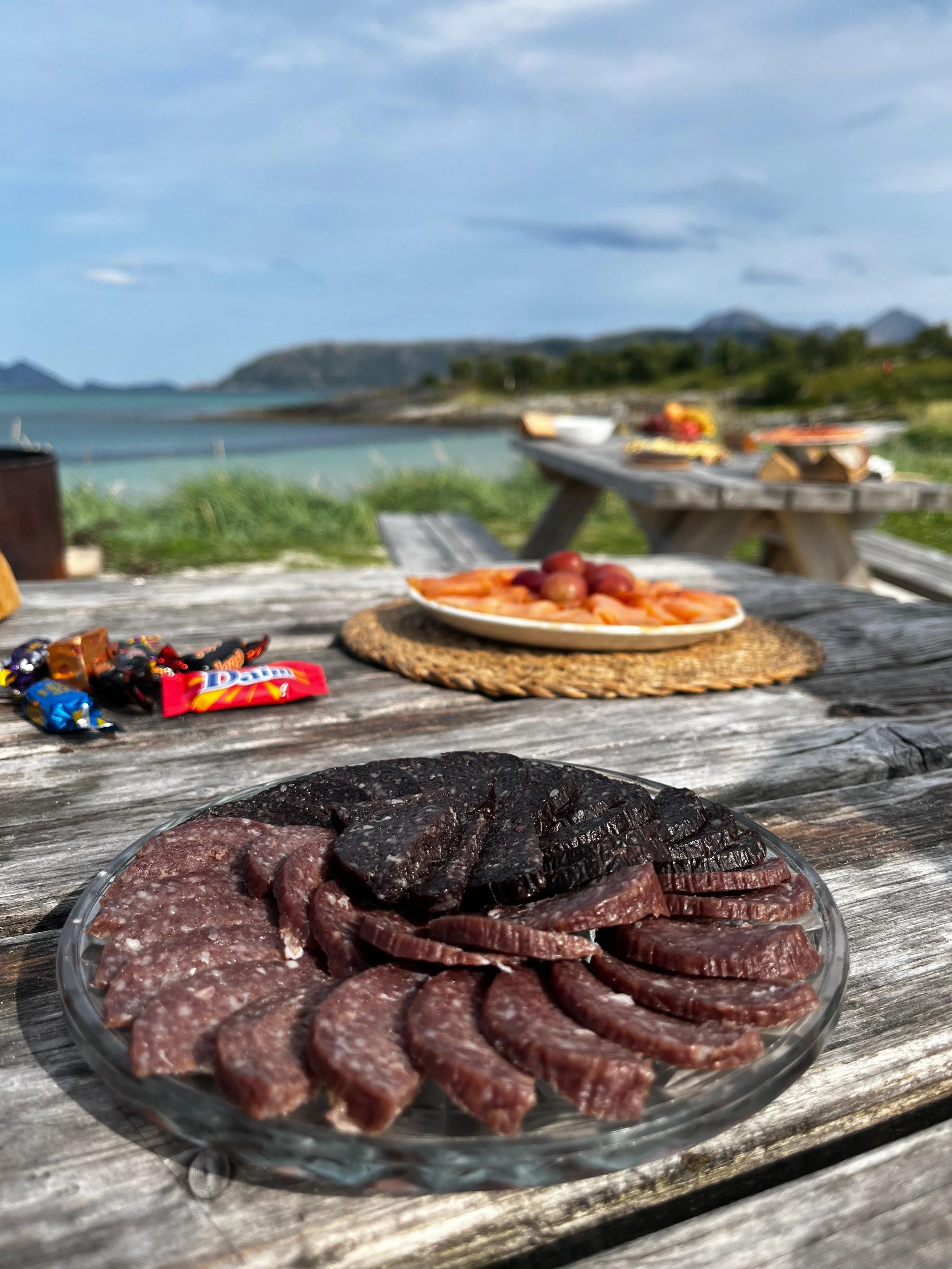 A picnic table with a plate of sliced cured meats, a plate of grapes and sliced fruit, and assorted candies, set outdoors near a body of water with boats and hills in the background.