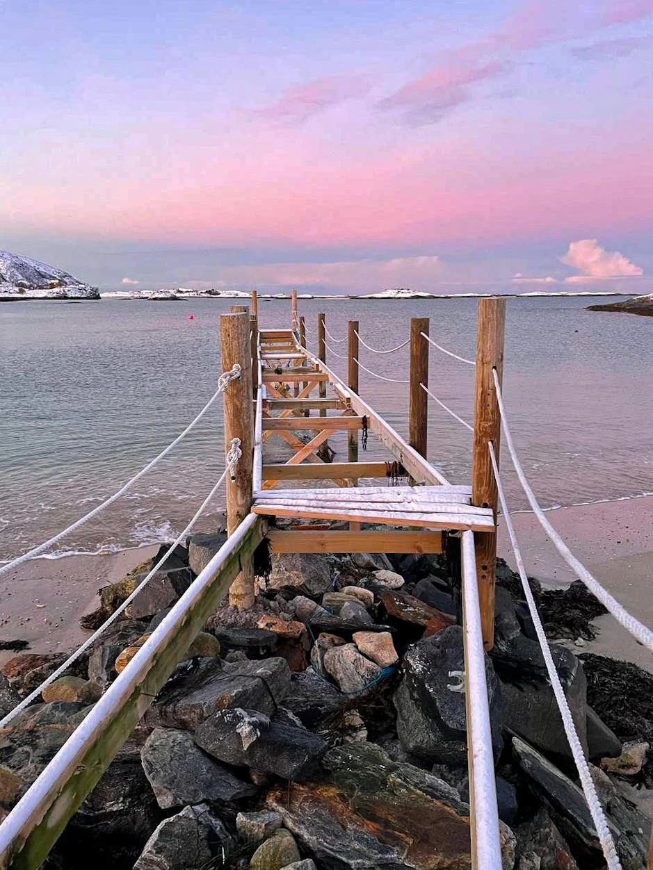 A wooden dock with snow on the planks extends over rocky shore into calm water, with a pink and purple sky and snow-covered mountains in the background.