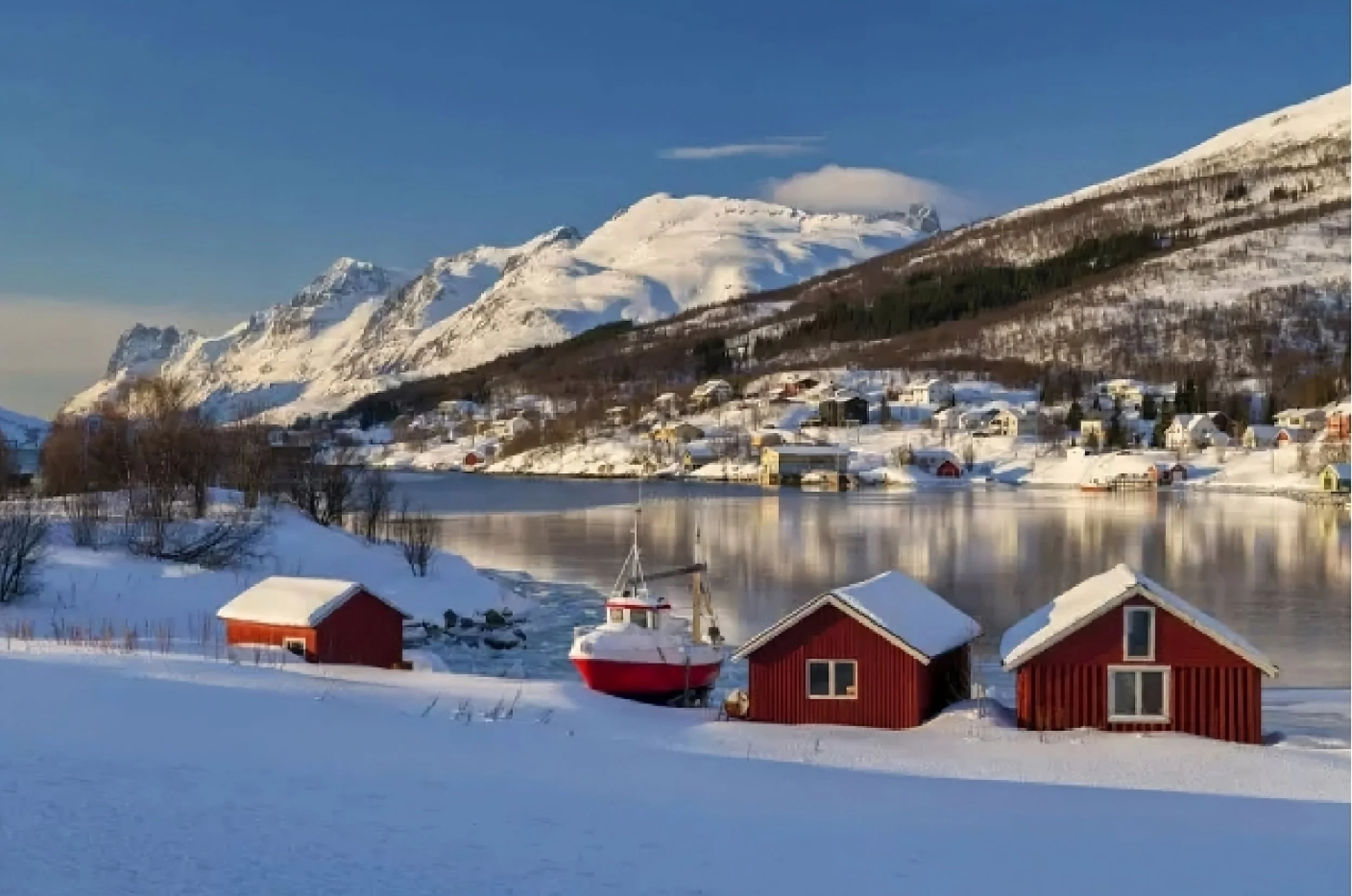 Snow-covered houses and boats along a waterfront, with snow-capped mountains in the background under a clear blue sky.