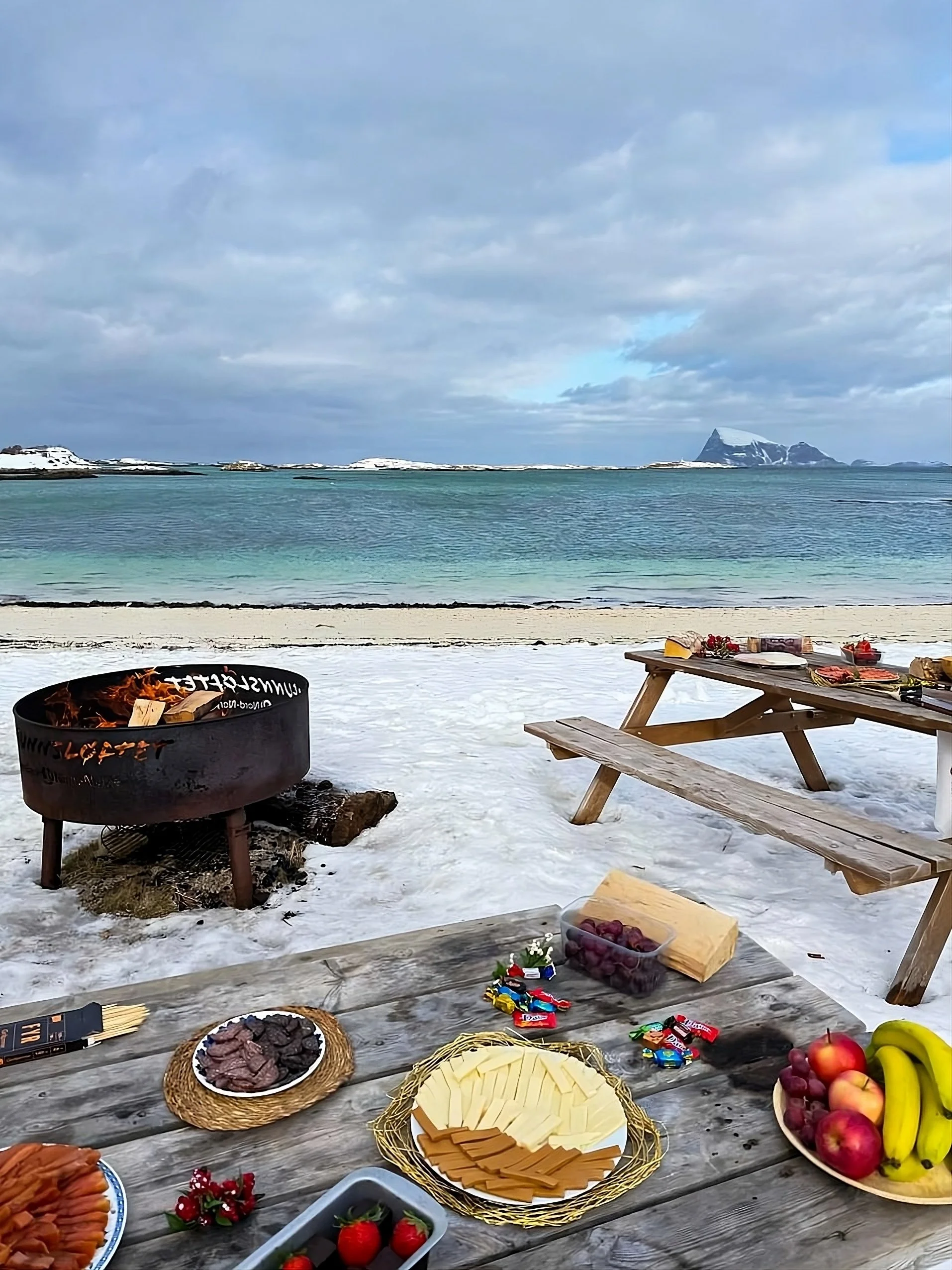 Beach picnic setup with food on a wooden table, a fire pit, picnic benches, and an ocean view with mountains in the background.