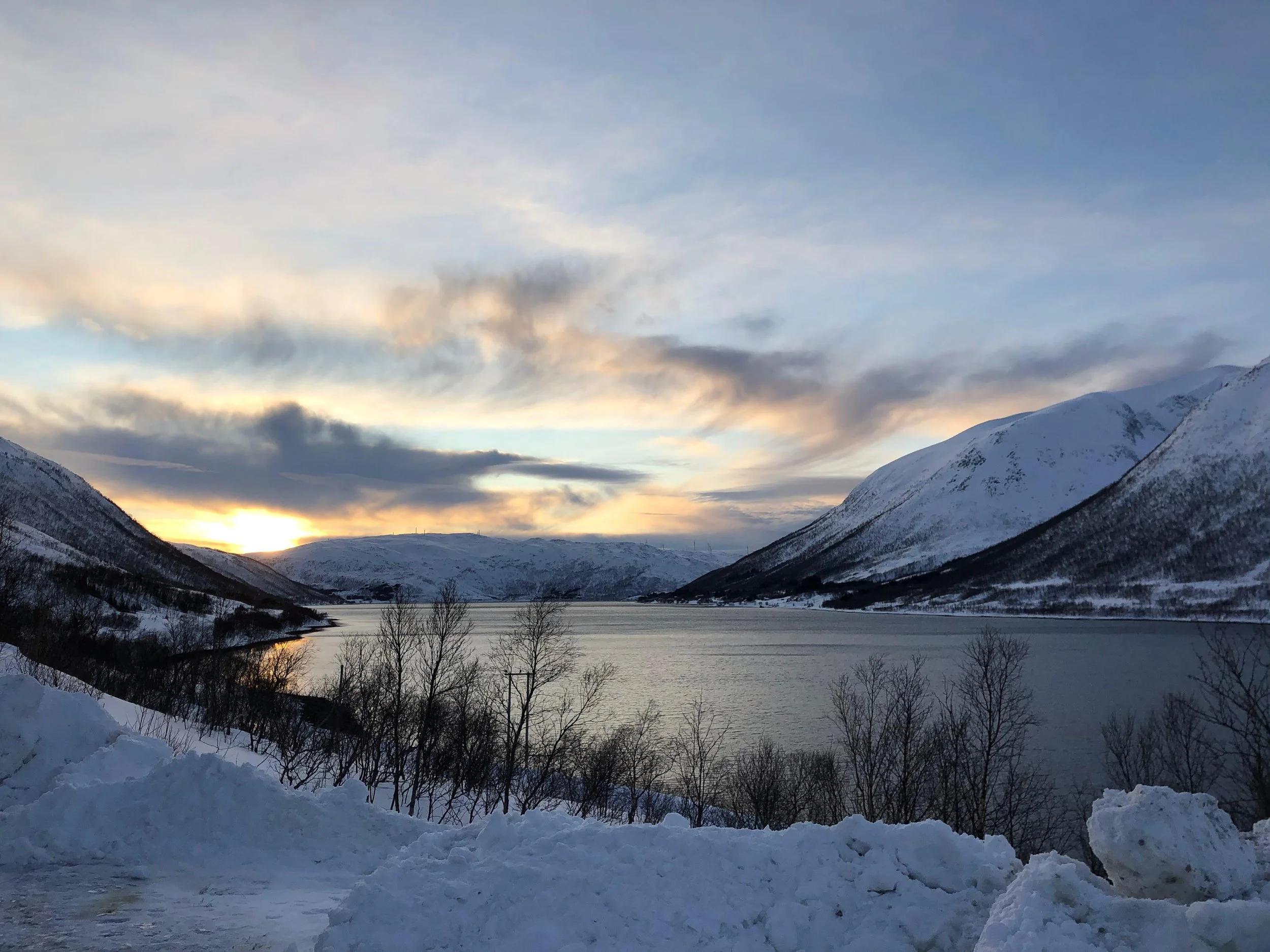 Snow-covered mountains surrounding a calm lake during sunset with a partly cloudy sky.