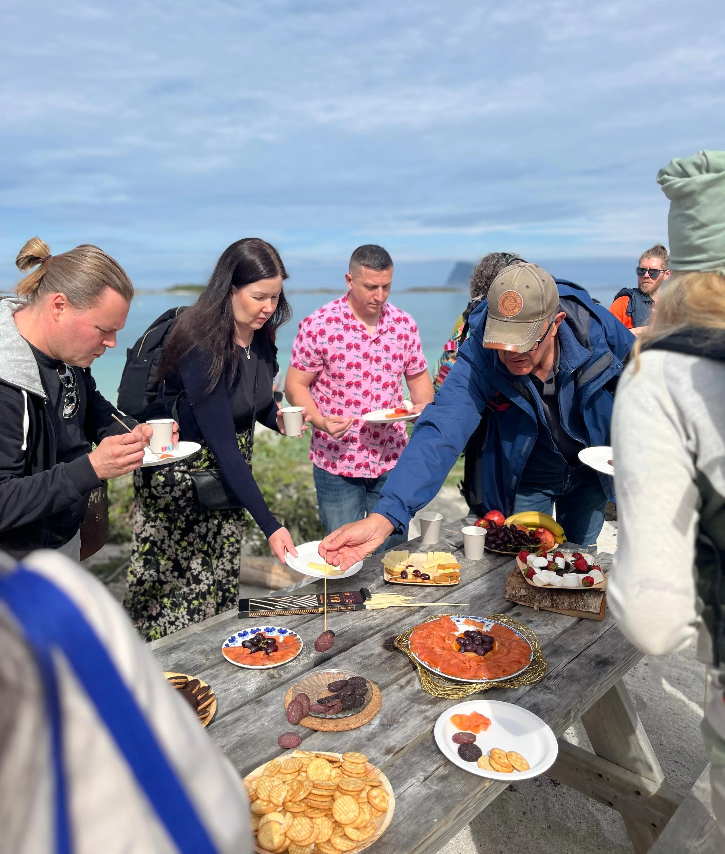 People gathered around a wooden table outdoors with various foods, including crackers, smoked salmon, cheese, crackers, berries, and fruit, during a social event near the beach with scenic ocean and sky background.