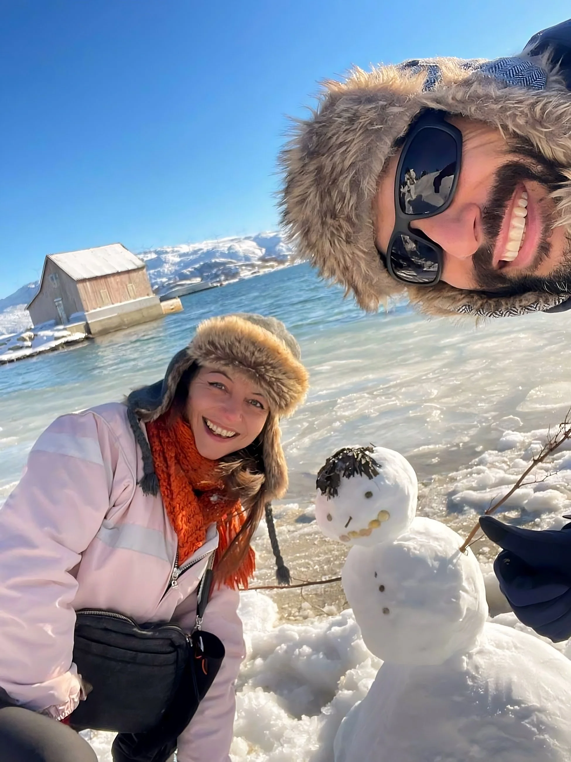 Two people dressed in winter clothing and sunglasses are smiling next to a small snowman on a snowy beach with snow-covered mountains and a blue sky in the background.