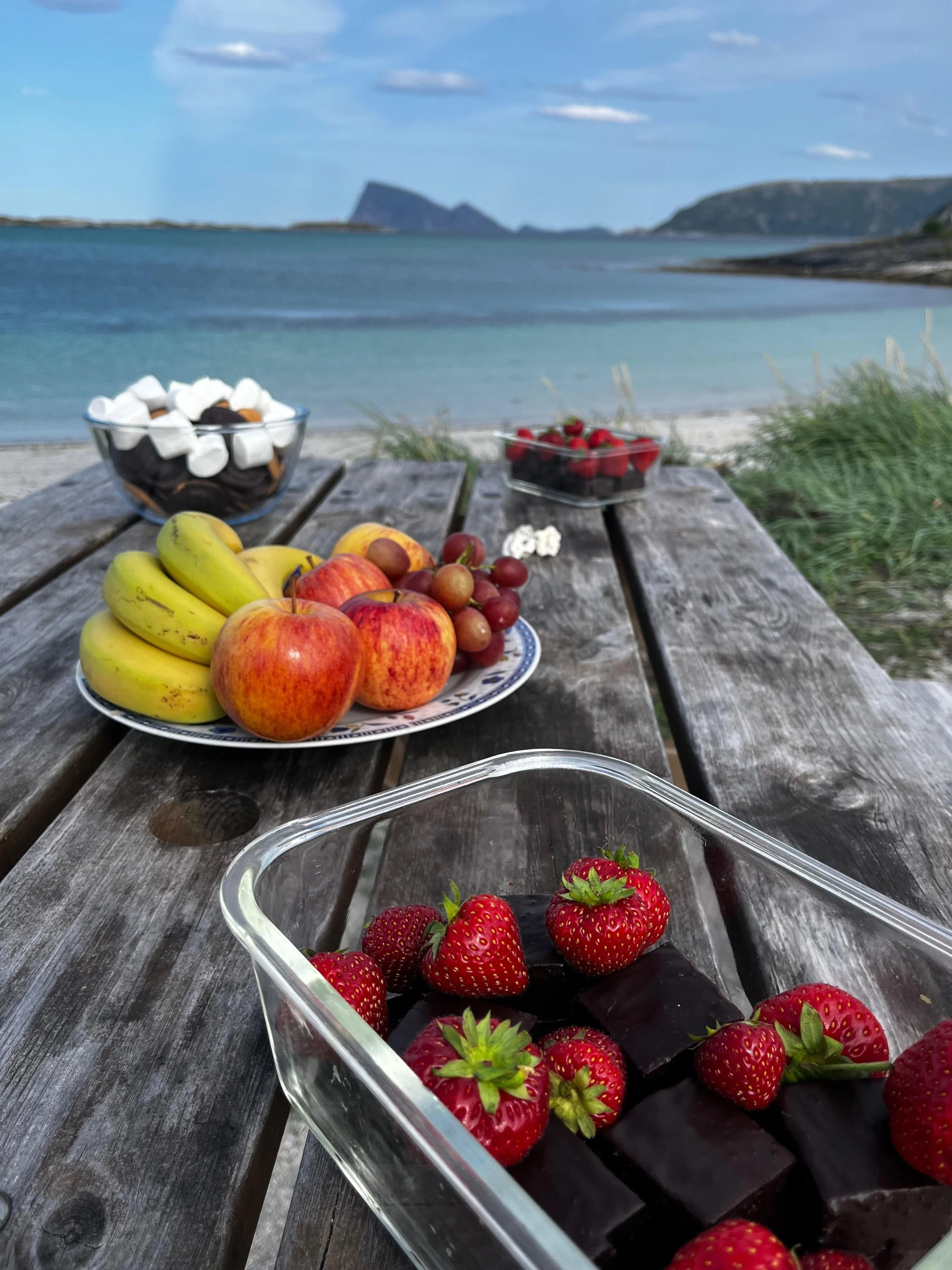 assortment of fresh fruit, including strawberries, apples, bananas, grapes, and chocolate snacks, arranged on a rustic wooden table by the beach with a view of the ocean and distant islands.