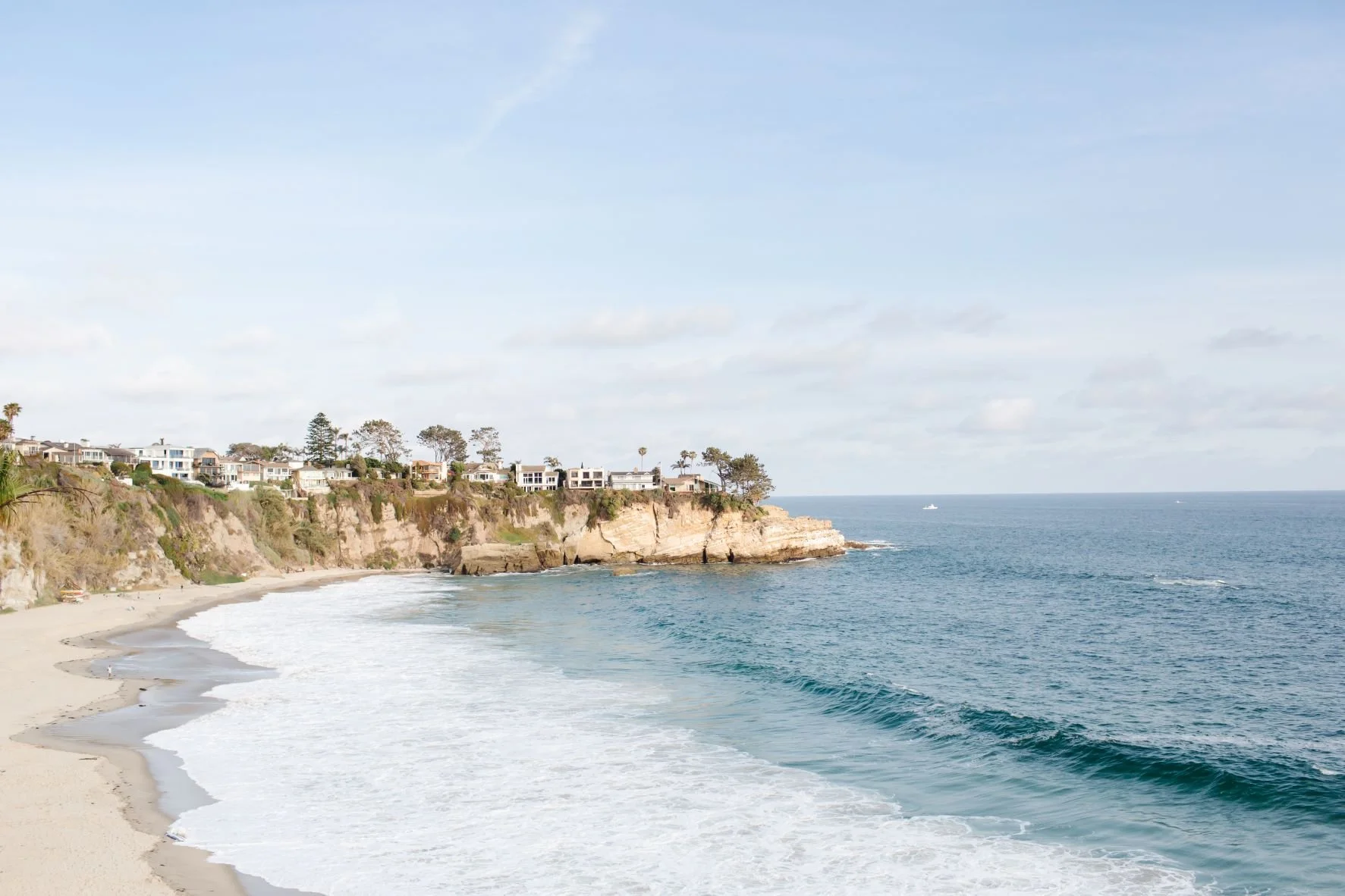 A sandy beach with waves crashing on the shore, rocky cliffside with houses on top, and the ocean stretching out to the horizon under a partly cloudy sky.
