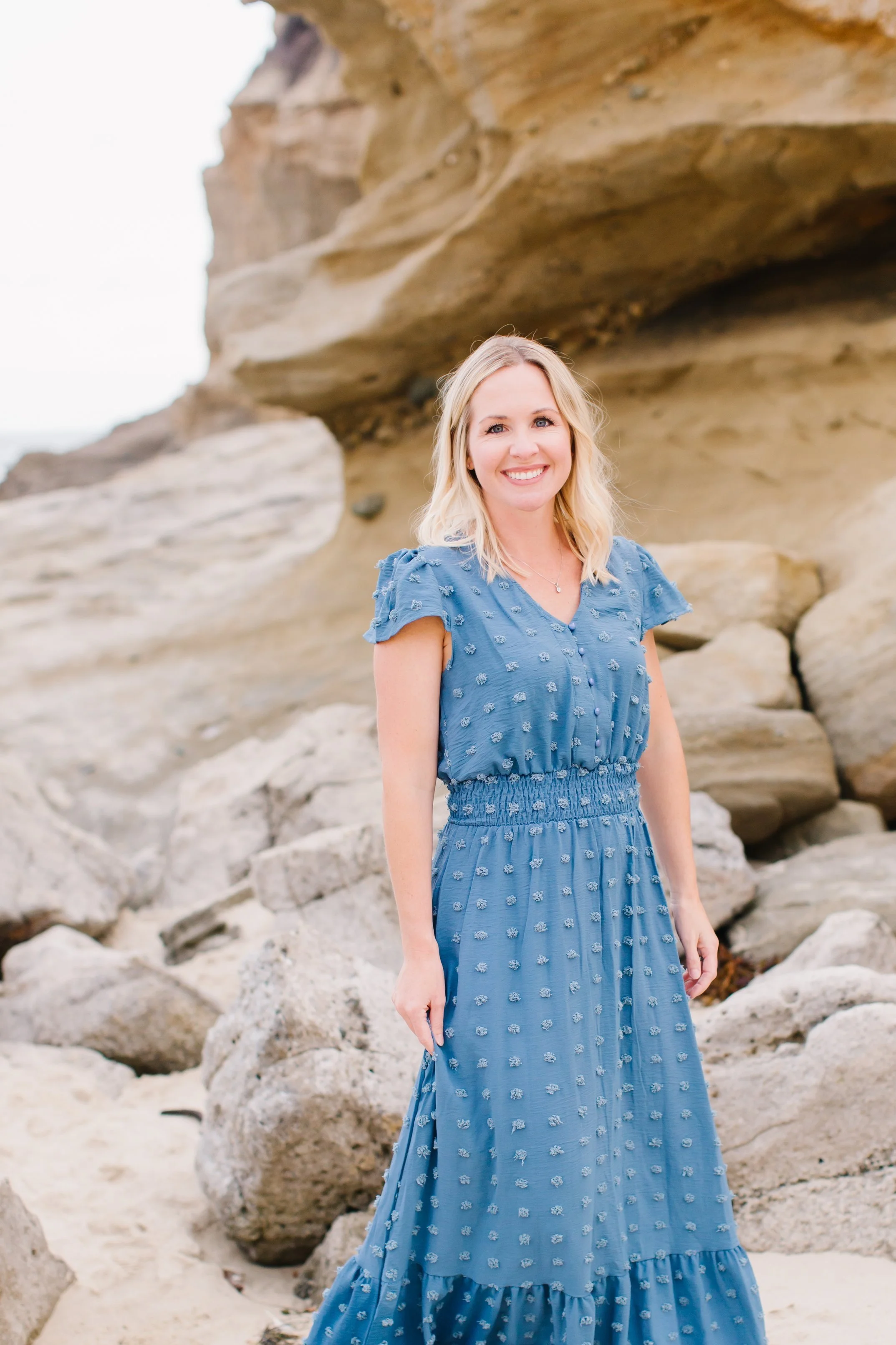 A woman with blonde hair wearing a blue dress stands on a rocky beach with large sandstone formations behind her.